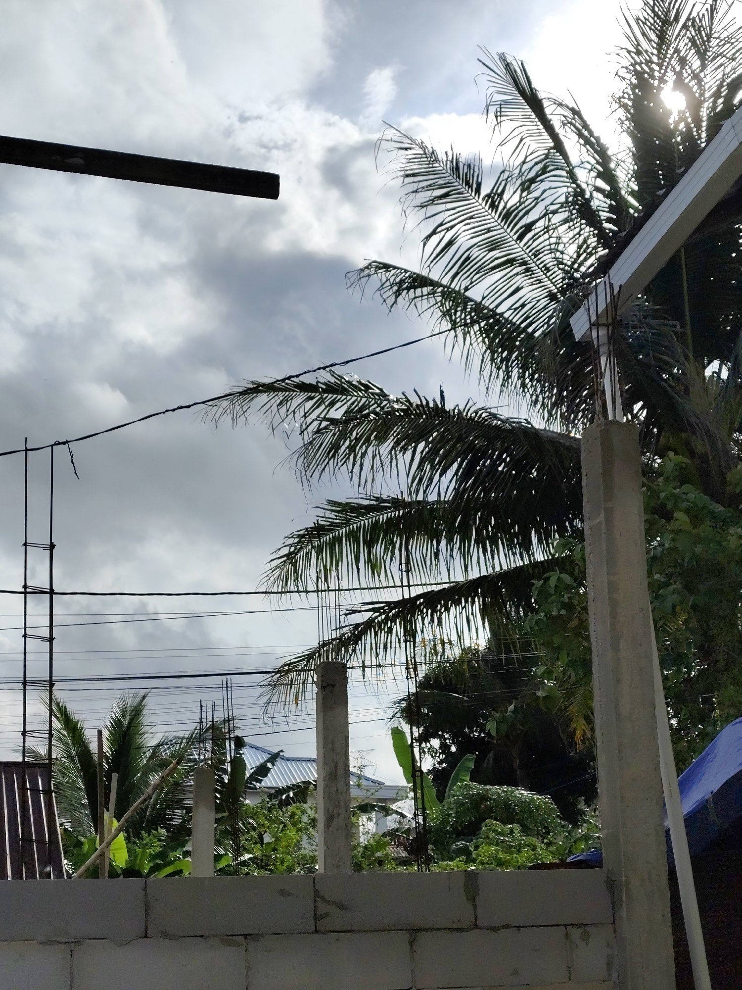 The image depicts a construction site with a cloudy sky and palm trees, suggestive of a tropical climate, likely in Balikpapan, Indonesia. In the foreground, a wall made of concrete blocks is visible, suggesting ongoing construction. Several concrete pillars and metal scaffolding are also present, indicating structural work. Overhead, power lines crisscross the sky, with palm fronds from nearby trees visible against the sky. The sun is partially obscured by clouds and palm leaves, casting a diffused light on the scene. In the background, parts of houses with corrugated metal roofs can be seen amidst lush greenery. The overall scene conveys a sense of development and everyday life in a tropical urban environment.