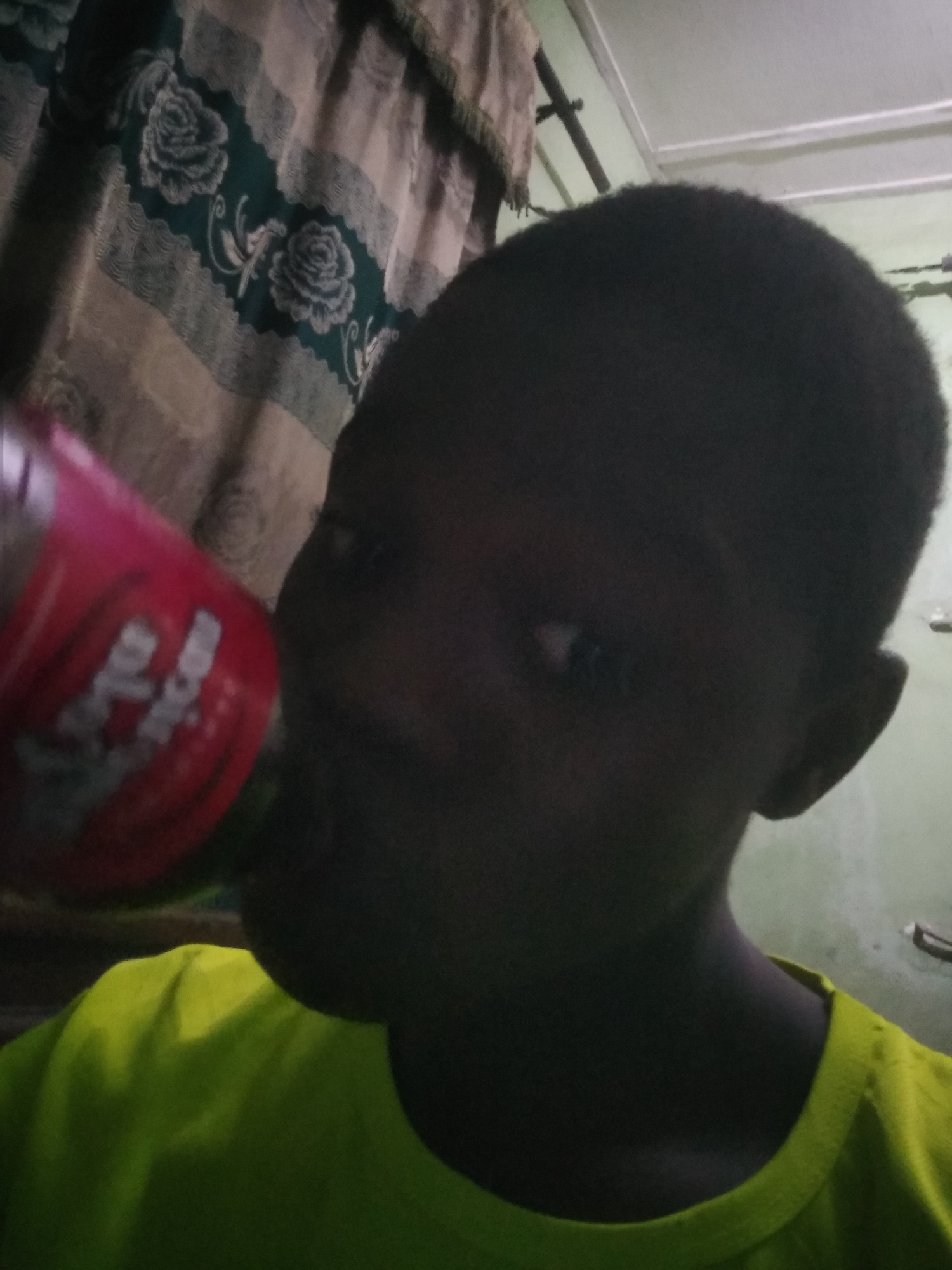 A young person of African descent is drinking from a red beverage container. The person is wearing a yellow-green shirt. The background includes patterned curtains and a light-colored wall. The location is unknown, and there is no information to suggest this is Shangisha, Nigeria.