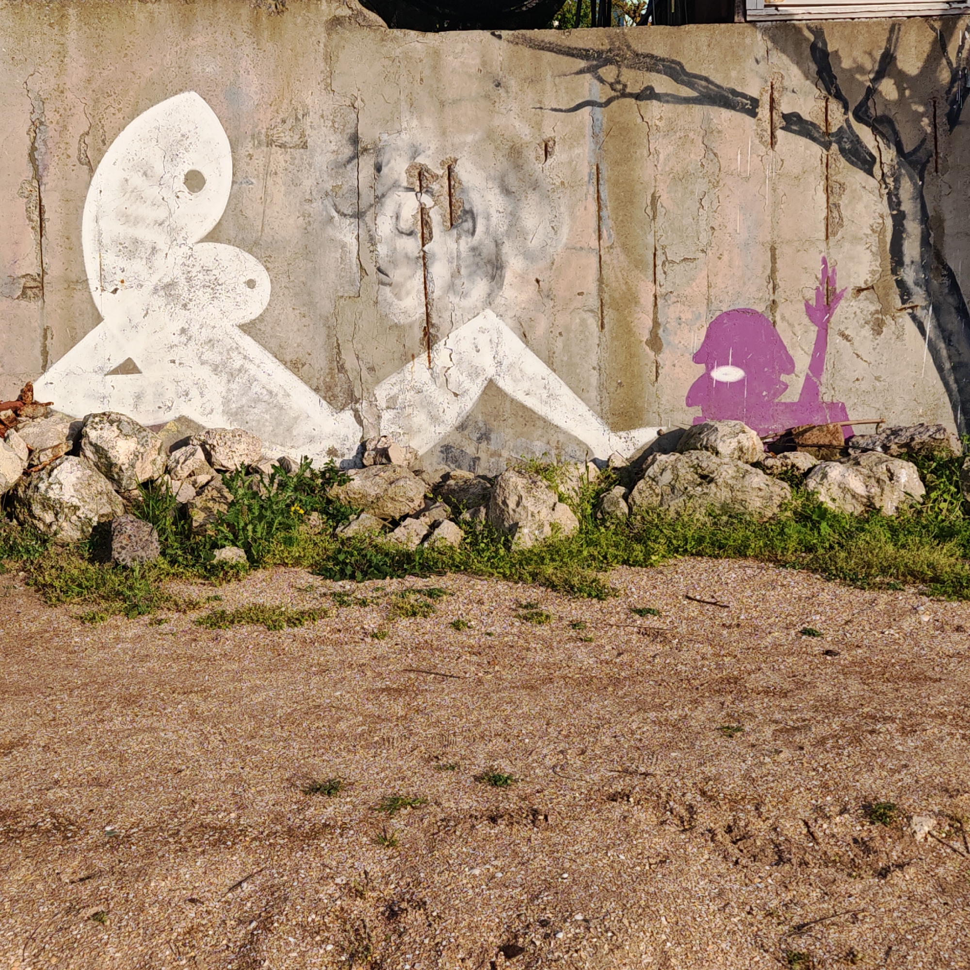 The image presents a concrete wall adorned with street art in an outdoor urban setting. On the wall, there's a white figure with a distinct circular feature at its head. Adjacent to it, a faint greyish portrait is visible, possibly faded or aged. Further to the right, a purple figure stands near a stylized tree branch. At the base of the wall, a pile of rocks is interspersed with small patches of green vegetation. The foreground features a coarse, brown ground. The location appears to be in the city of Unknown, Unknown.