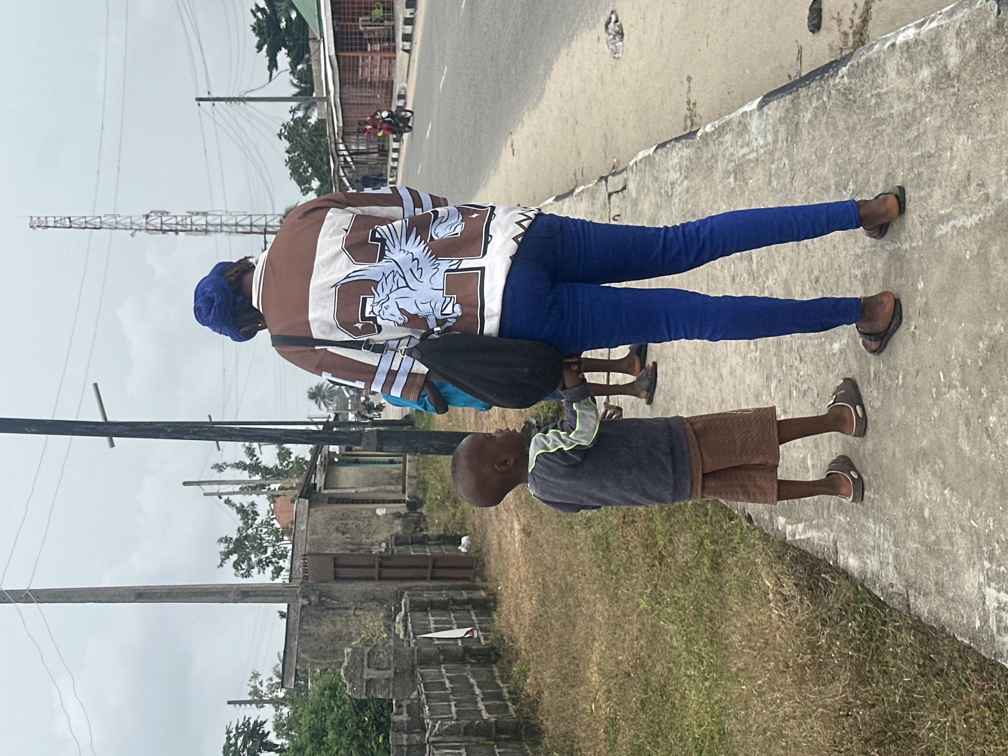 In Badagry, Nigeria, a young child stands on a concrete sidewalk next to a woman wearing a blue headscarf and a brown and white jacket with a Pegasus design. The child is wearing a gray long-sleeved shirt with neon green trim and brown shorts. The woman is wearing blue jeans and dark sandals. Behind them, a busy street with a single lane of traffic stretches into the distance. Buildings line the opposite side of the street, and power lines crisscross the sky. A cell tower is visible on the horizon. The sidewalk is bordered by dry, brown grass.