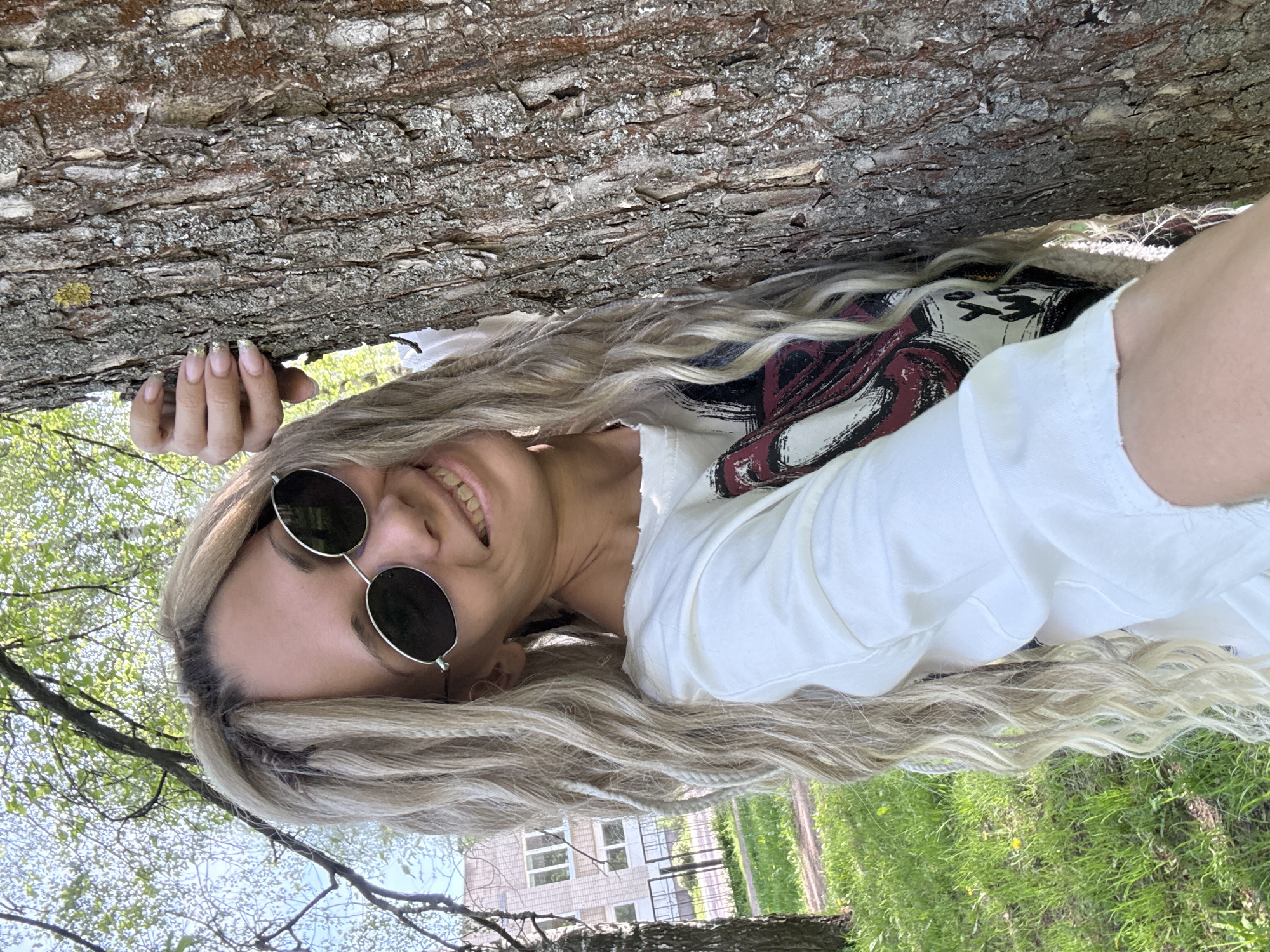 The image depicts a woman with long, wavy blonde hair and sunglasses lying under a tree. She is wearing a white shirt with a design and holding onto the tree trunk with her hand. The background shows green grass, trees, and a building, suggesting an outdoor location in a park or green space. The location is Rusanovo, Russia.