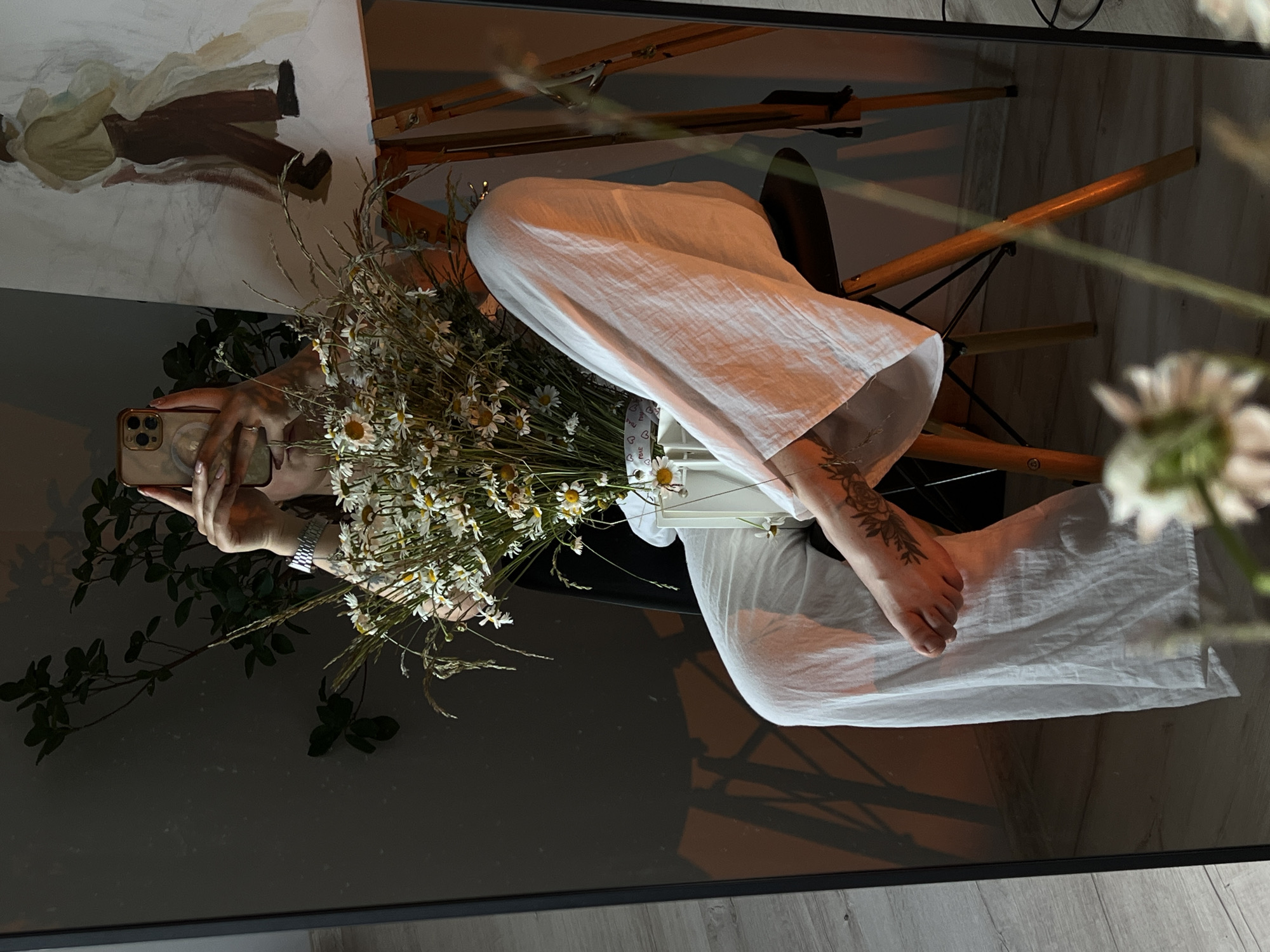 In an indoor setting, a person is captured in a reflection within a large, black-framed mirror. The person, seated on a black chair with wooden legs, holds a smartphone to take a selfie. They are wearing loose-fitting white pants. A bouquet of white daisies and grasses is held in front of them, resting in a white vase. A painting is visible in the background, displayed on an easel. The floor appears to be light-colored wood. Shadows from the chair and possibly plants are cast on the dark wall behind the person. The overall lighting suggests an evening or dimly lit environment, with warm tones visible on the right side of the frame, likely from artificial light. The scene is devoid of any specific identifiable landmarks of Ternopil, Ukraine.
