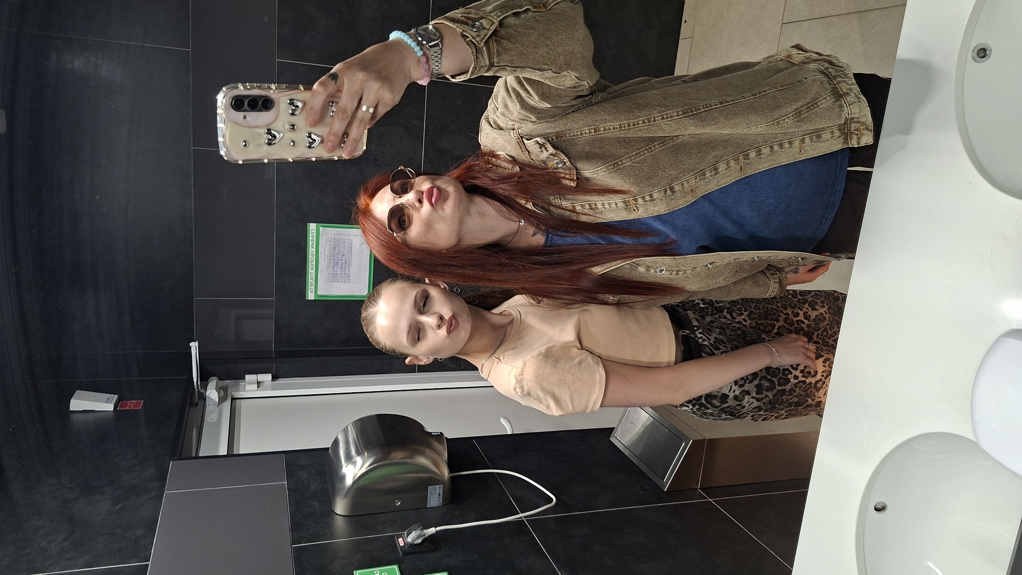 In a well-lit public restroom with dark tiled walls, two individuals are taking a mirror selfie. On the left, a young woman with blonde hair pulled back in a ponytail wears a beige t-shirt and a leopard-print skirt. She is winking and pursing her lips, looking towards the camera. To her right, an adult woman with long, reddish-brown hair and sunglasses is wearing a denim jacket over a blue t-shirt. She is also pursing her lips, with her right hand holding up a smartphone with a heart-patterned case. Visible in the background are a hand dryer mounted on the wall, a trash can, a door, and what appears to be a sign with text. The scene is set on a white countertop with sinks visible. The location is identified as Асіпавіцкі сельскі Савет, Belarus.