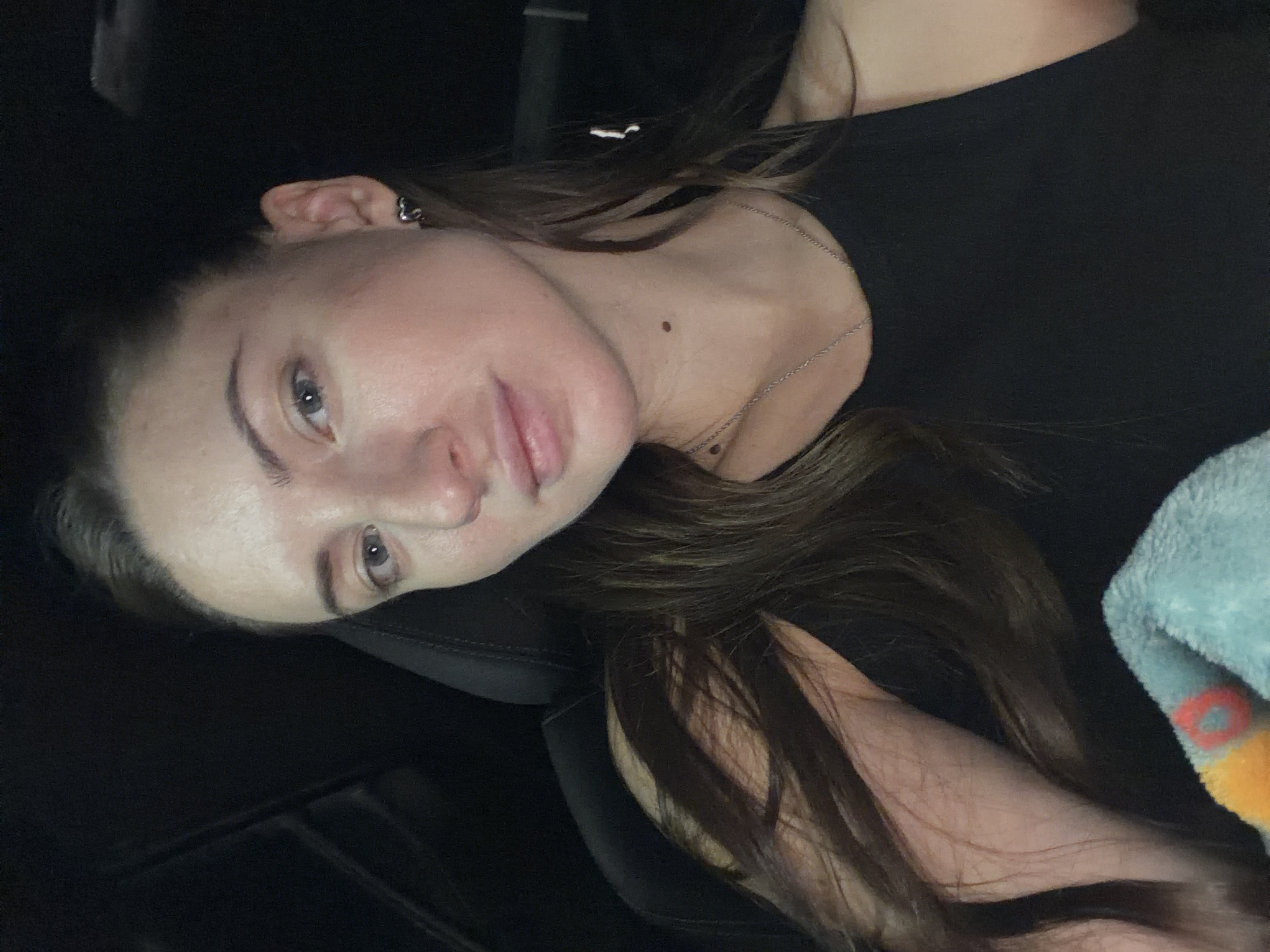 A young woman with long, dark brown hair, fair skin, and blue eyes is captured in a close-up, selfie-style photograph. She is wearing a black tank top and a silver chain necklace. A small, heart-shaped earring is visible in her left ear. Her hair is parted on the left and falls over her left shoulder. She appears to be in a dimly lit environment, possibly the interior of a car, with dark upholstery visible behind her. A portion of a light blue and orange patterned blanket is visible in the lower left corner of the frame. The photograph does not contain any elements that specifically indicate a location in Sharm El Sheikh, Egypt.