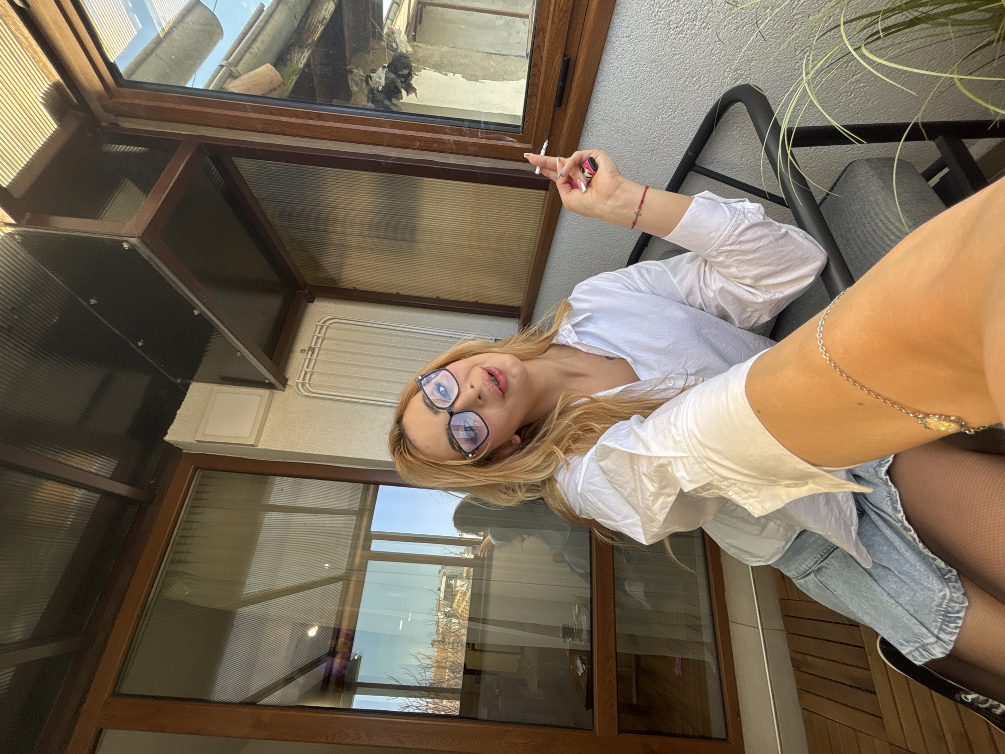 A woman is sitting in a chair on a balcony or patio, holding a lit cigarette. She is wearing glasses, a white collared shirt, a blue jean skirt, and black tights. Her blond hair is long and loose. The location appears to be an enclosed balcony or patio with windows. Outside the window, there is a view of urban rooftops and architectural details. The lighting suggests it is daytime. The exact location in Varna, Bulgaria, cannot be determined from the image alone.