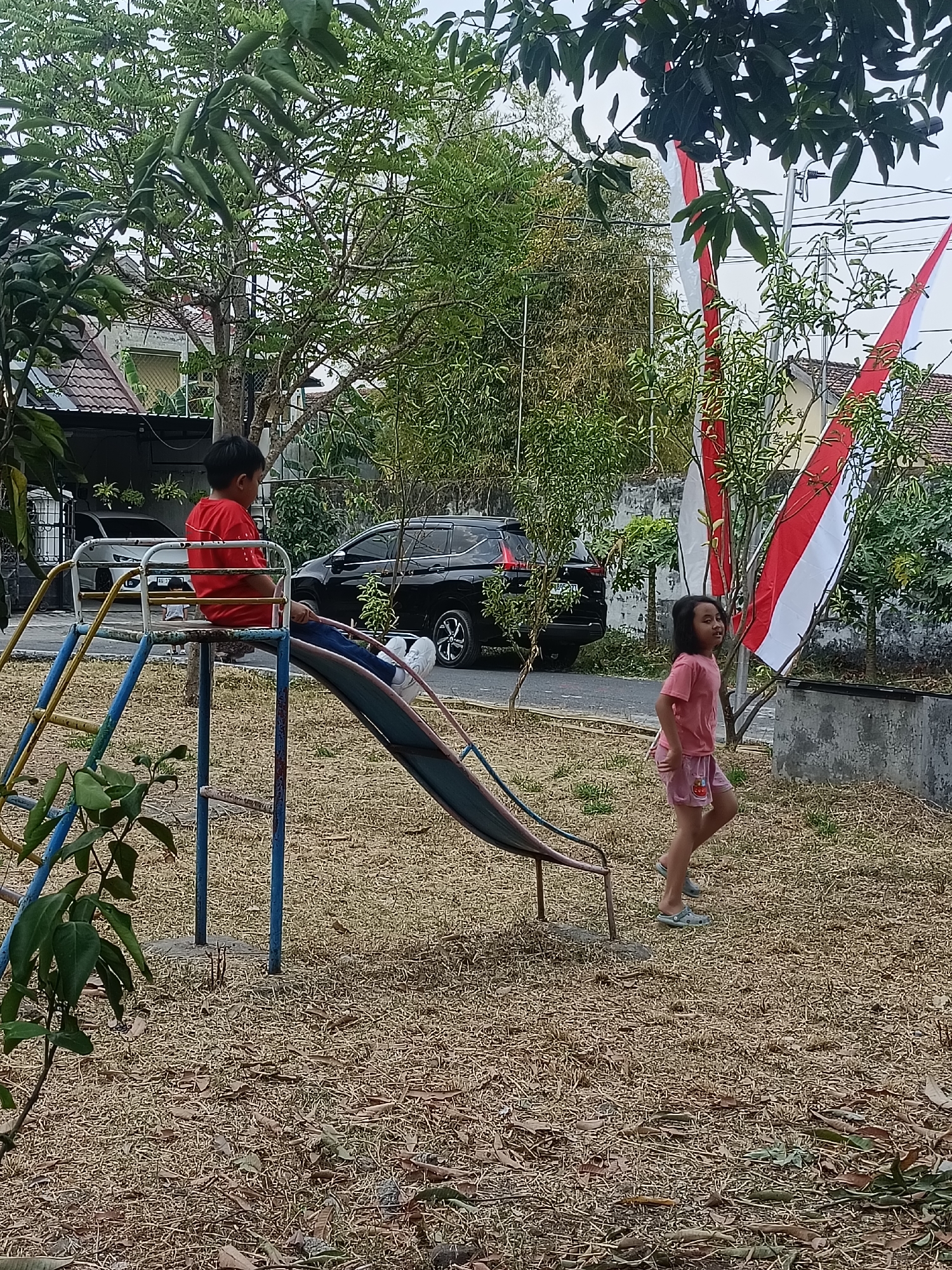 The image depicts a playground scene in Kediri City, Indonesia, with two children present. On the left, a young boy wearing a red t-shirt and blue jeans is sitting on a metal slide, looking downwards. To the right of the slide, a young girl in a pink t-shirt and pink shorts is walking away from the viewer, looking over her shoulder. The playground features a multi-colored metal slide with blue poles and a yellow ladder. The ground is covered with dry grass and fallen leaves. In the background, there are trees, houses, and two cars parked on a paved road. Two Indonesian flags, red and white, are visible hanging from poles amidst the foliage. The overall environment suggests an outdoor setting in a residential area.