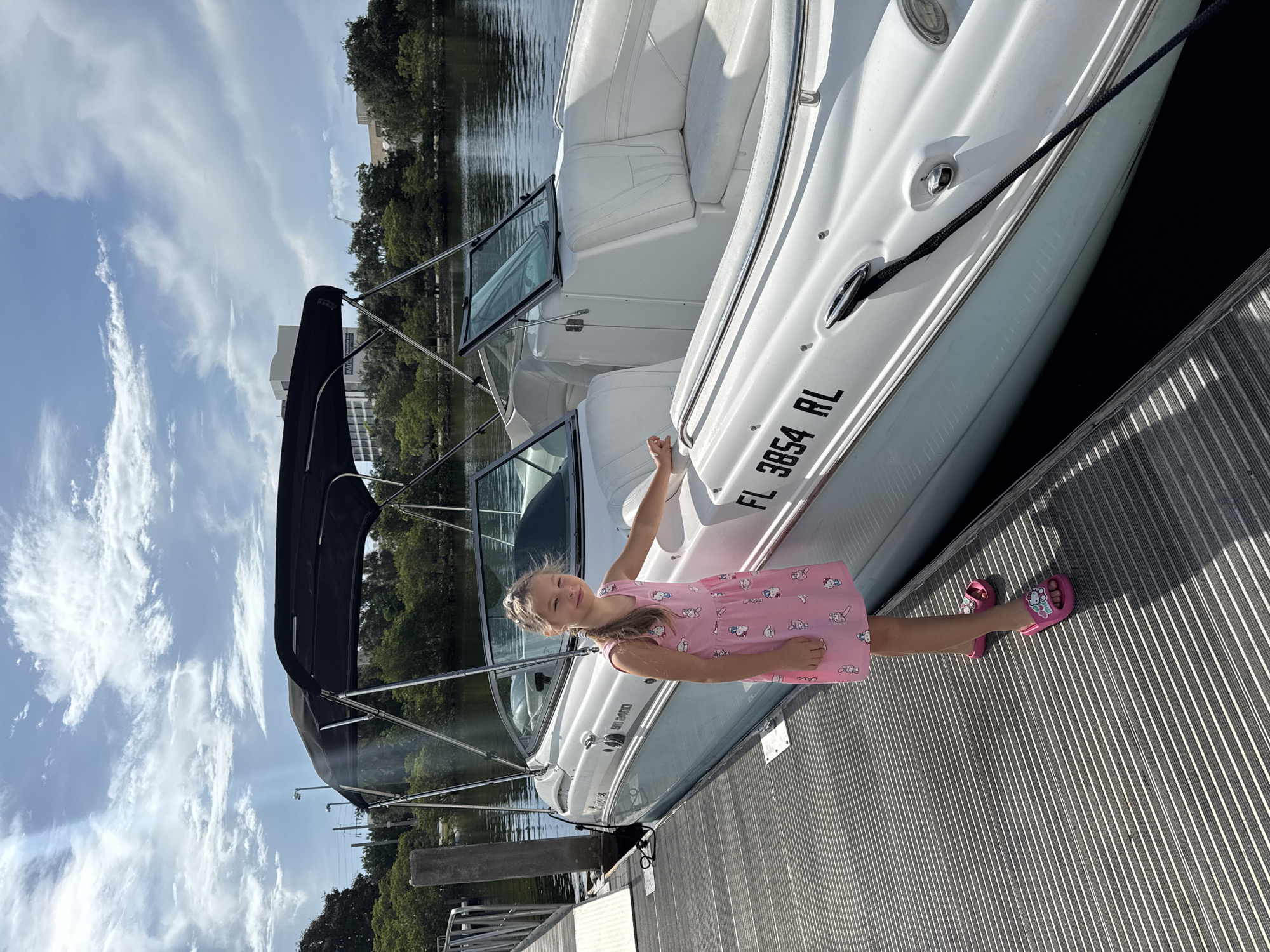 A young girl with blonde hair, wearing a pink dress adorned with cartoon cat figures and pink slide sandals, stands on a wooden dock next to a white boat. The boat has "FL 3854 RL" printed on its side. The girl is smiling and looking towards the camera. The scene appears to be a sunny day, indicated by the bright lighting and the blue sky with scattered white clouds. The boat is docked on a body of water, likely a canal or river, with trees and buildings visible in the background, suggesting an urban or suburban waterside setting in Fort Lauderdale, USA. The girl's pose and smile convey a sense of happiness and enjoyment, likely on a recreational outing.