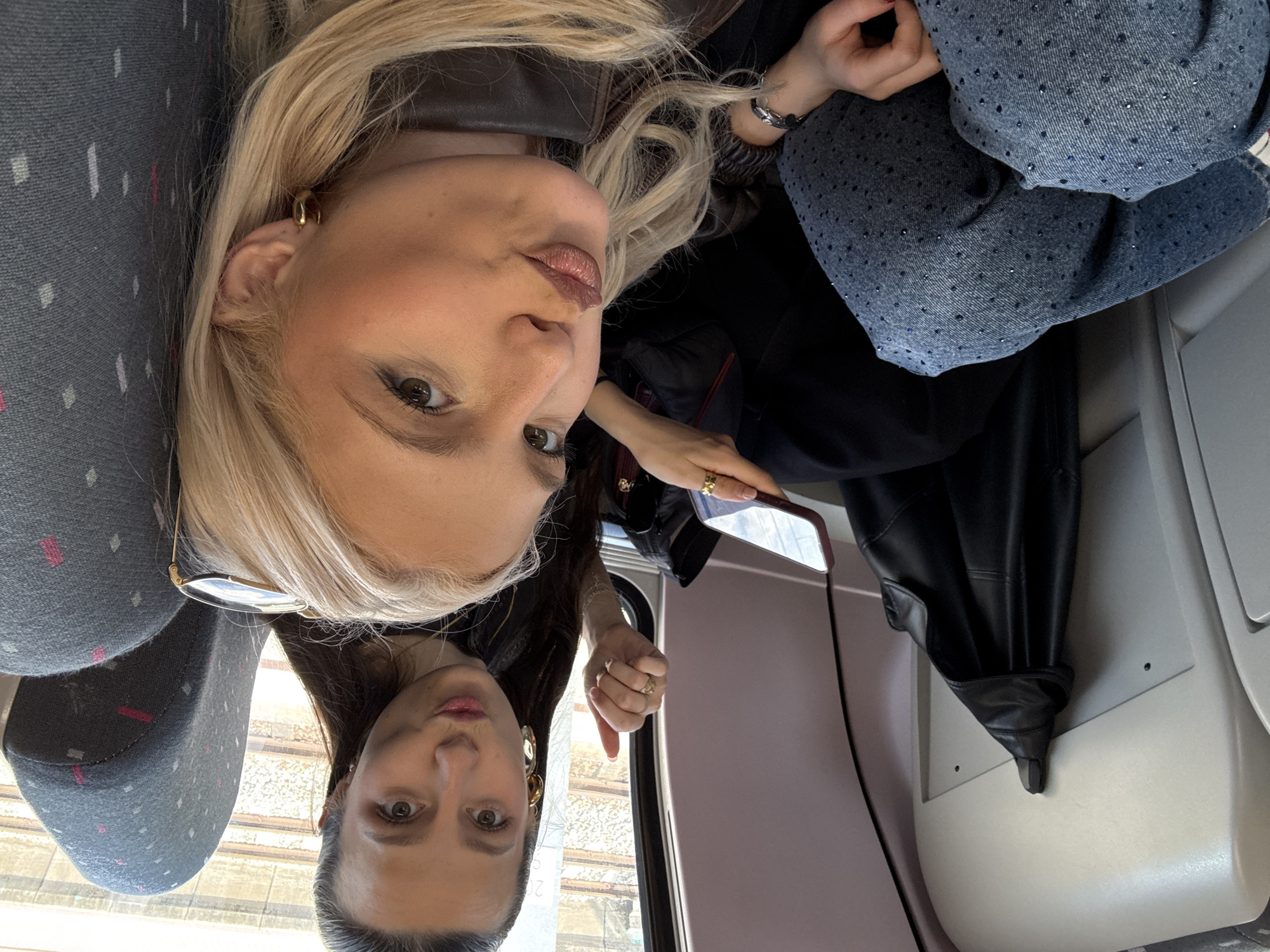 Two women are taking a selfie on what appears to be a train. The woman in the foreground has blond hair, is wearing hoop earrings, a brown leather jacket and a bracelet. The woman in the background has dark hair and is wearing large drop earrings. They are seated on a train with grey seats. The background shows a view of train tracks. In the background, a black leather jacket is hanging on a hook.
It's not possible to determine the exact location within Vilvoorde, Belgium, based on the image alone. More information would be required to pinpoint the specific train line or area.