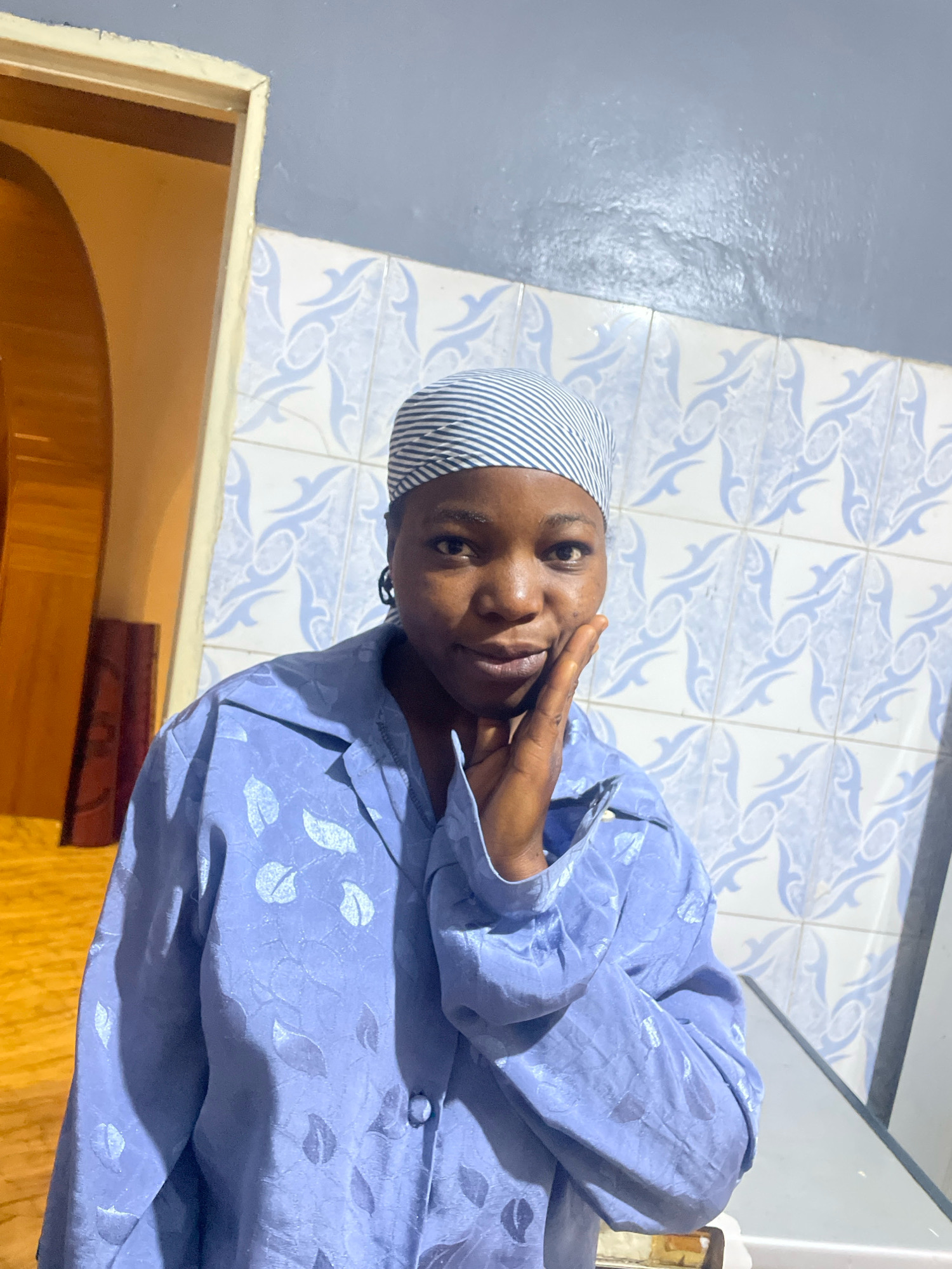 A young Black woman wearing a blue patterned shirt and a striped headscarf stands indoors, her right hand resting on her cheek. She faces the camera with a neutral expression. The background features blue and white patterned tiles on the wall and a doorway to her left, which opens into a dimly lit space with a wooden arch. The scene appears to be an interior residential setting within the city of Jos, Nigeria.