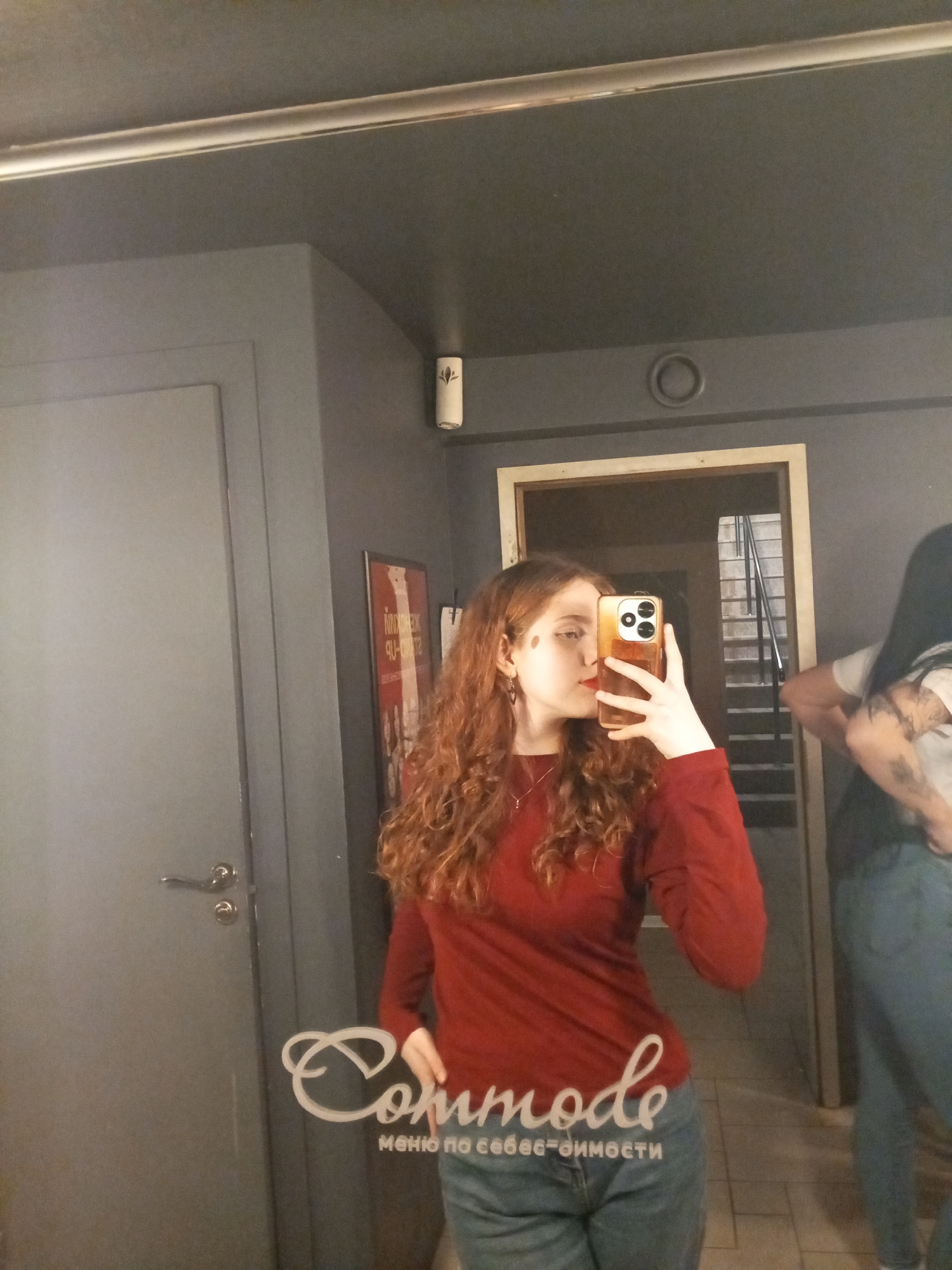 A fair-skinned woman with curly red hair and a red lipstick is taking a selfie in a mirror using a gold-colored smartphone. She's wearing a long-sleeved red shirt and blue jeans. In the background, partially visible on the right, another person is standing, wearing a white shirt, blue jeans, and has long dark hair. They have tattoos on their arm.
The location appears to be a bathroom or hallway with gray walls and a silver trim near the ceiling. A door with a silver handle is visible on the left. There's a poster on the wall behind the woman. The text "Commode" is superimposed on the lower part of the image, with text in Russian underneath it that reads "меню по себе-имость". The location is in Saint Petersburg, Russia, based on the signage and the language used. In the mirror's reflection, a staircase is also visible.