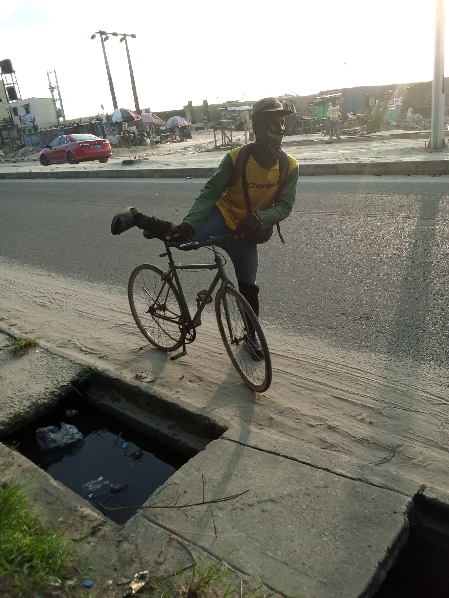 A person stands beside a bicycle on a concrete sidewalk next to a street in Ado, Nigeria. The person is wearing a green long-sleeved shirt over a yellow shirt with "Chowdek" printed on it, jeans, and a hat. The face is partially covered. A leg with a boot is draped over the bicycle handlebars. To the left is a concrete storm drain filled with dark, possibly stagnant water and trash. Behind the person is a street with a red car, pedestrians, and buildings under a bright, possibly sunny sky. Utility poles are also visible.