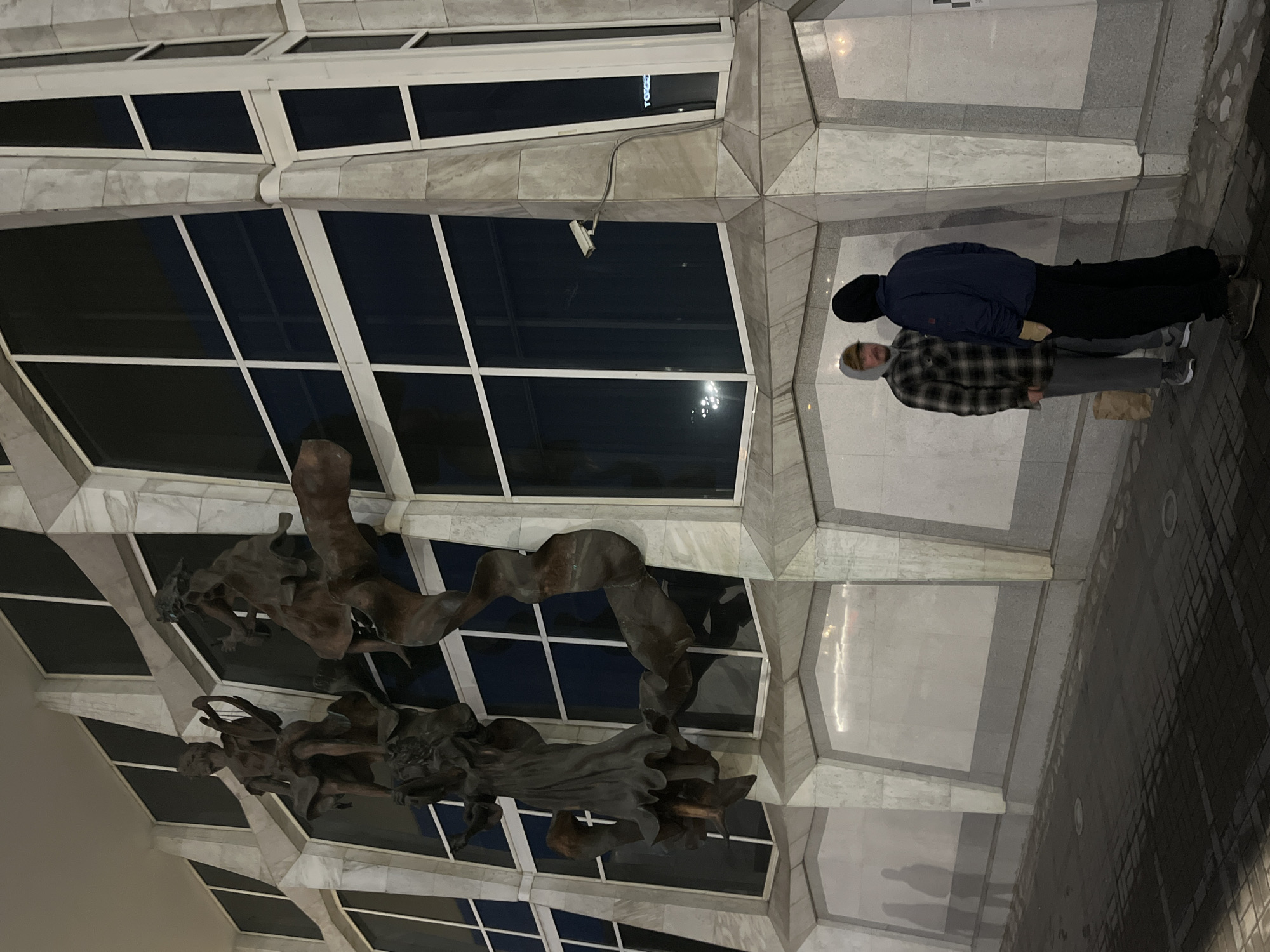 The image depicts two individuals standing on a gray tile surface beneath a modern architectural structure. The building features a grid-like pattern of windows and marble-like panels, with a bronze-colored sculpture hanging from the ceiling. A security camera is visible near the building’s corner. The individuals are dressed in casual clothing. The location is Krasnoyarsk, Russia, based on the architecture and sculpture style which match locations in that city.