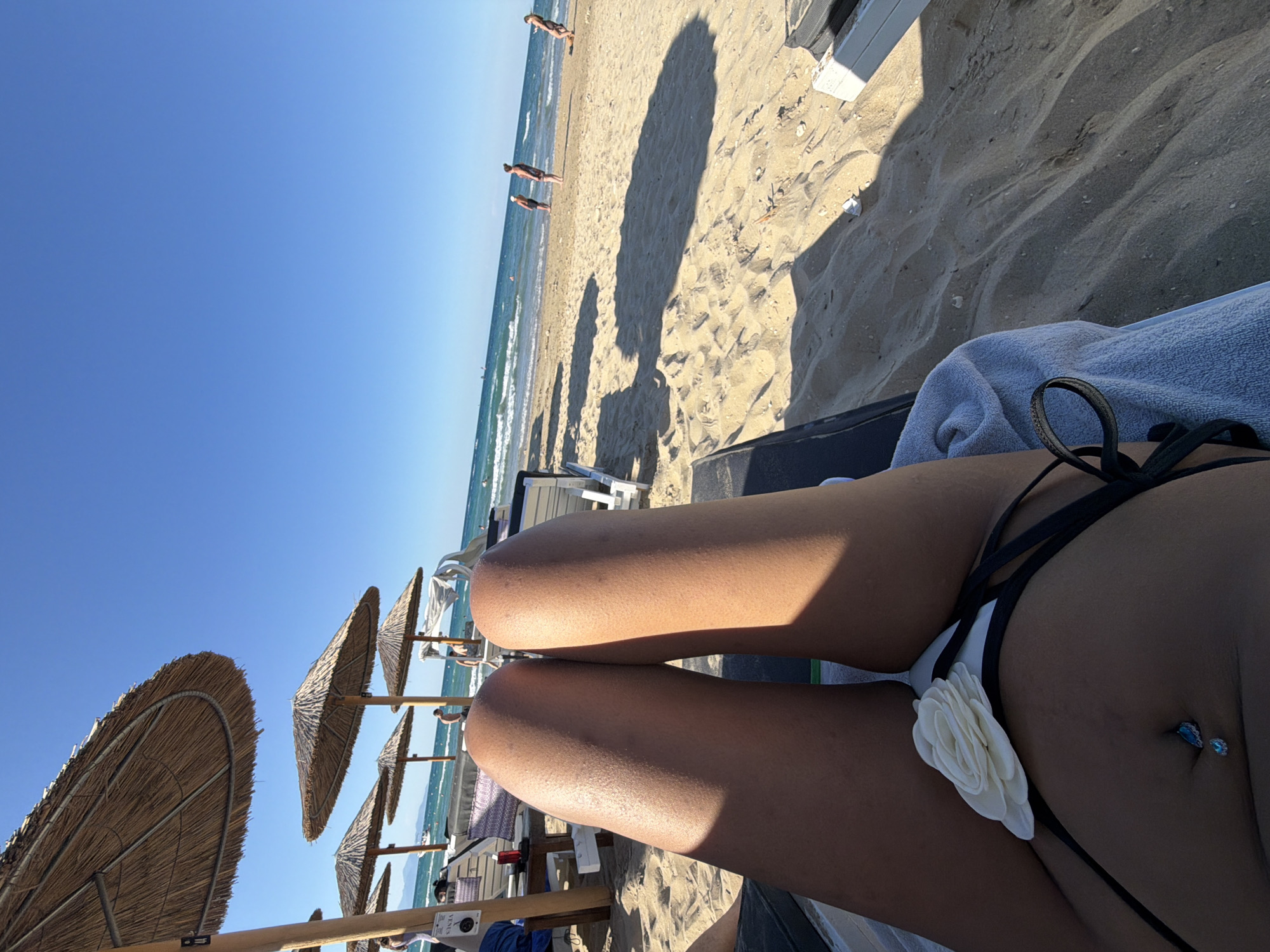 In the city of Arkadii Municipal Unit, Greece, an individual lies on a beach chair facing away from the ocean. Their tanned legs, adorned with a white flower detail on their bikini bottoms, are positioned in the foreground. The beach sand stretches out before them, dotted with other beach chairs and patrons enjoying the sunny weather. In the background, the turquoise waters of the Aegean Sea meet the horizon under a clear blue sky. A few people can be seen walking along the shore or wading in the water. Beach umbrellas provide shade on the sandy expanse.