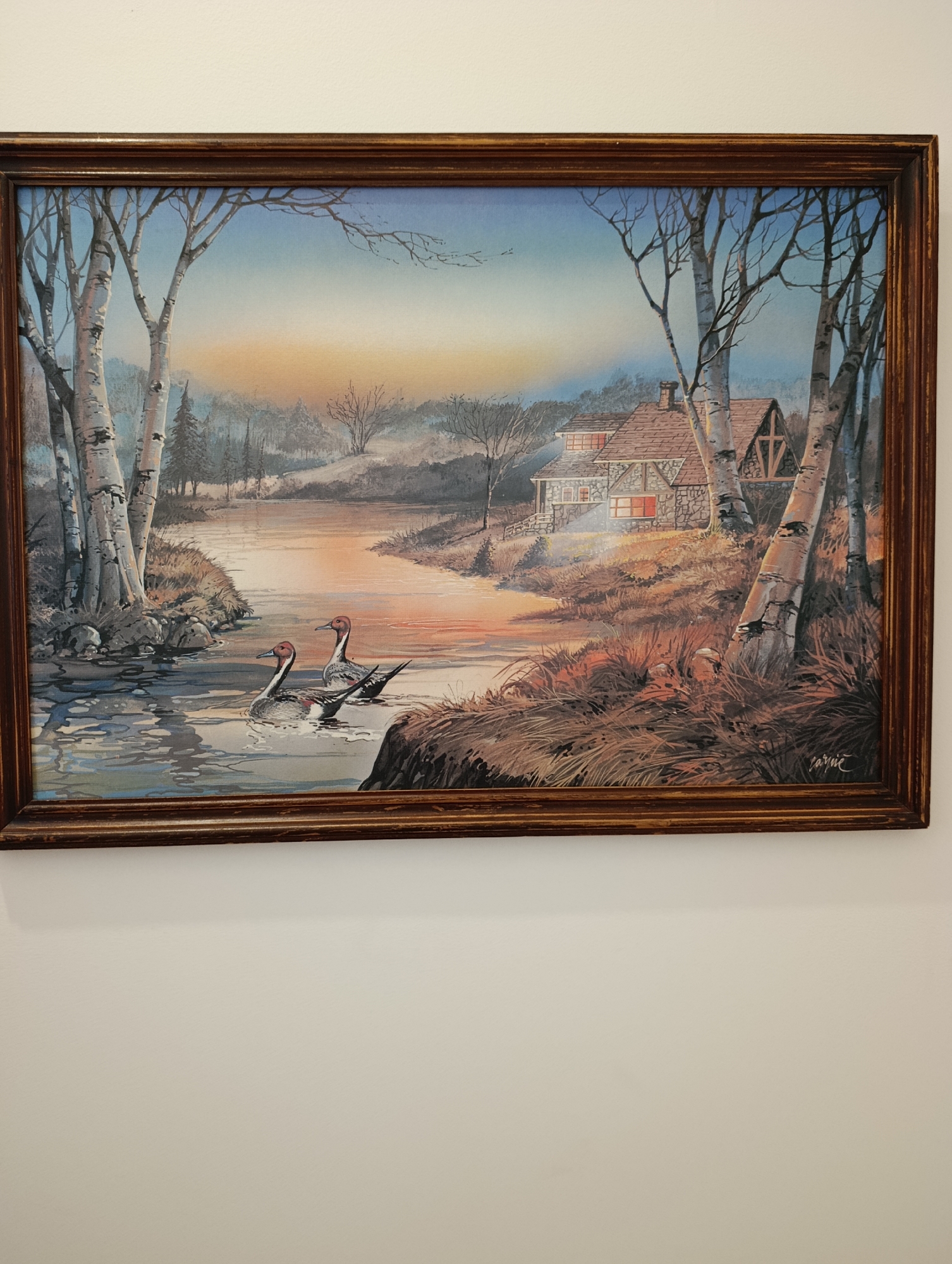 This is a framed painting of a serene landscape. The scene depicts a tranquil river winding through a wooded area. On the left bank, several birch trees with white bark and dark markings are prominent. The river flows gently, with two ducks swimming in the foreground. The right bank of the river is lined with tall, dry grass and some rocky outcroppings. Further in the background, a rustic stone house with a reddish-brown tiled roof is visible, with warm light emanating from its windows, suggesting it is dusk or dawn. The sky is a gradient of blue at the top, transitioning to a soft orange and yellow near the horizon, indicating either sunrise or sunset. Bare trees with thin branches are scattered throughout the landscape, adding to the sense of a cool season, possibly autumn or early spring. The painting is framed by a dark, wooden frame with some visible texture. The signature "Jarnic" is visible in the bottom right corner of the painting. This image does not depict any specific location in the city of Setúbal, Portugal.