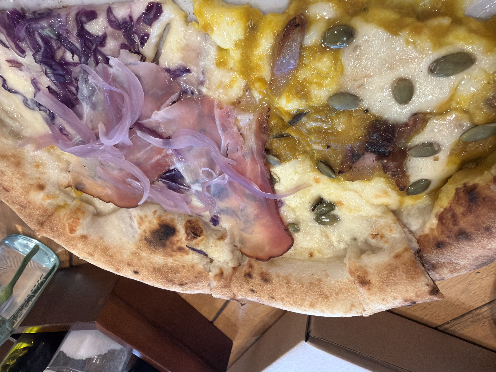 A close-up, high-angle shot depicts a pizza slice on a surface, presumably a table. The pizza features two distinct topping variations divided along a line. One side is topped with shredded red cabbage, translucent sliced red onions, and thin slices of prosciutto. The other side includes a yellow sauce, possibly pumpkin or butternut squash, scattered with green pumpkin seeds and mozzarella cheese. The crust is blistered and golden-brown. To the left, a portion of a clear glass jar or container with a green object inside is visible. Nearby are a wooden object and two transparent containers, one empty and the other potentially containing white granular material. The environment suggests an indoor dining setting. I have no information about this image being from Nha Trang Ward, Vietnam.