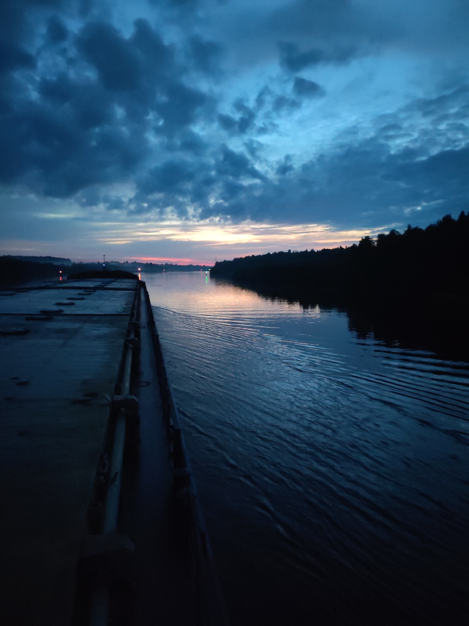 The image depicts a twilight scene over a wide river, possibly during dawn or dusk. Dark, ominous clouds dominate the upper portion of the sky, with streaks of pale orange and pink near the horizon, suggesting the last light of day or the first light of dawn. The water's surface is calm, reflecting the muted colors of the sky and creating gentle ripples from movement.
On the left side of the frame, a long, flat deck, likely part of a barge or a stationary structure, occupies a significant portion of the foreground. The deck appears to be made of concrete or a similar material and shows signs of wear. Several metal fixtures and railings are visible along the edge of the deck.
The right side of the image features a densely wooded shoreline, silhouetted against the sky. The trees are dark and indistinct due to the low light conditions. In the distance, along the river, several navigational lights are visible, indicated by small, colored points of light—red and green—emerging from the darkness. These lights suggest the presence of buoys or other markers in the waterway.
There are no people visible in the image. The overall atmosphere is serene, yet with a touch of somberness due to the dark clouds and the twilight. The location is identified as Bessonovo, Russia, implying this is a scene on a river in or near that region.