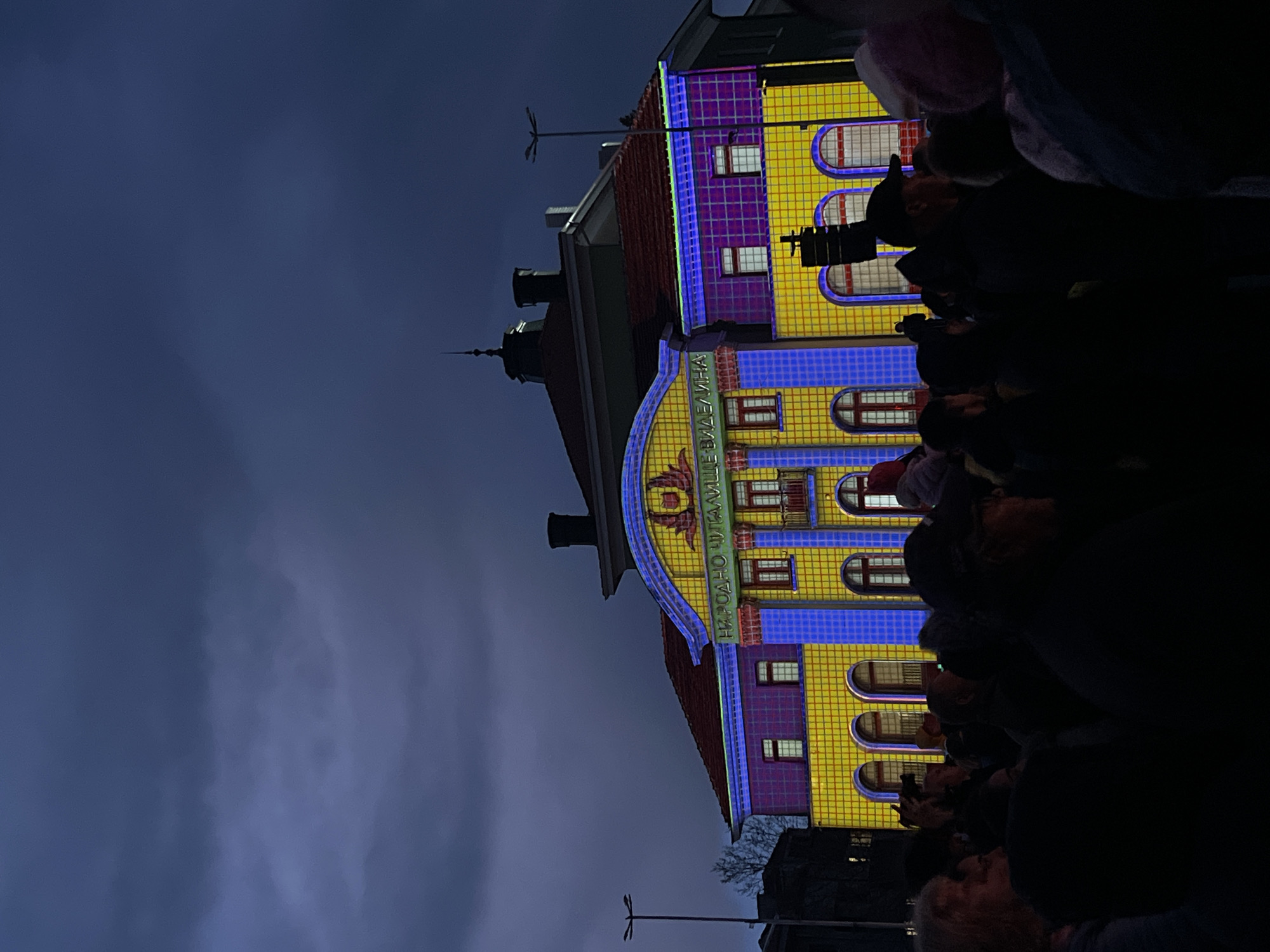 The image shows the facade of the Narodno Chitalishte (Community Cultural Center) "Videlina" in Panagyurishte, Bulgaria. The building is illuminated with yellow, purple, and blue lights, giving it a grid-like pattern. In the foreground, there is a crowd of people. The sky is a dark blue. There are streetlights in front of the building.