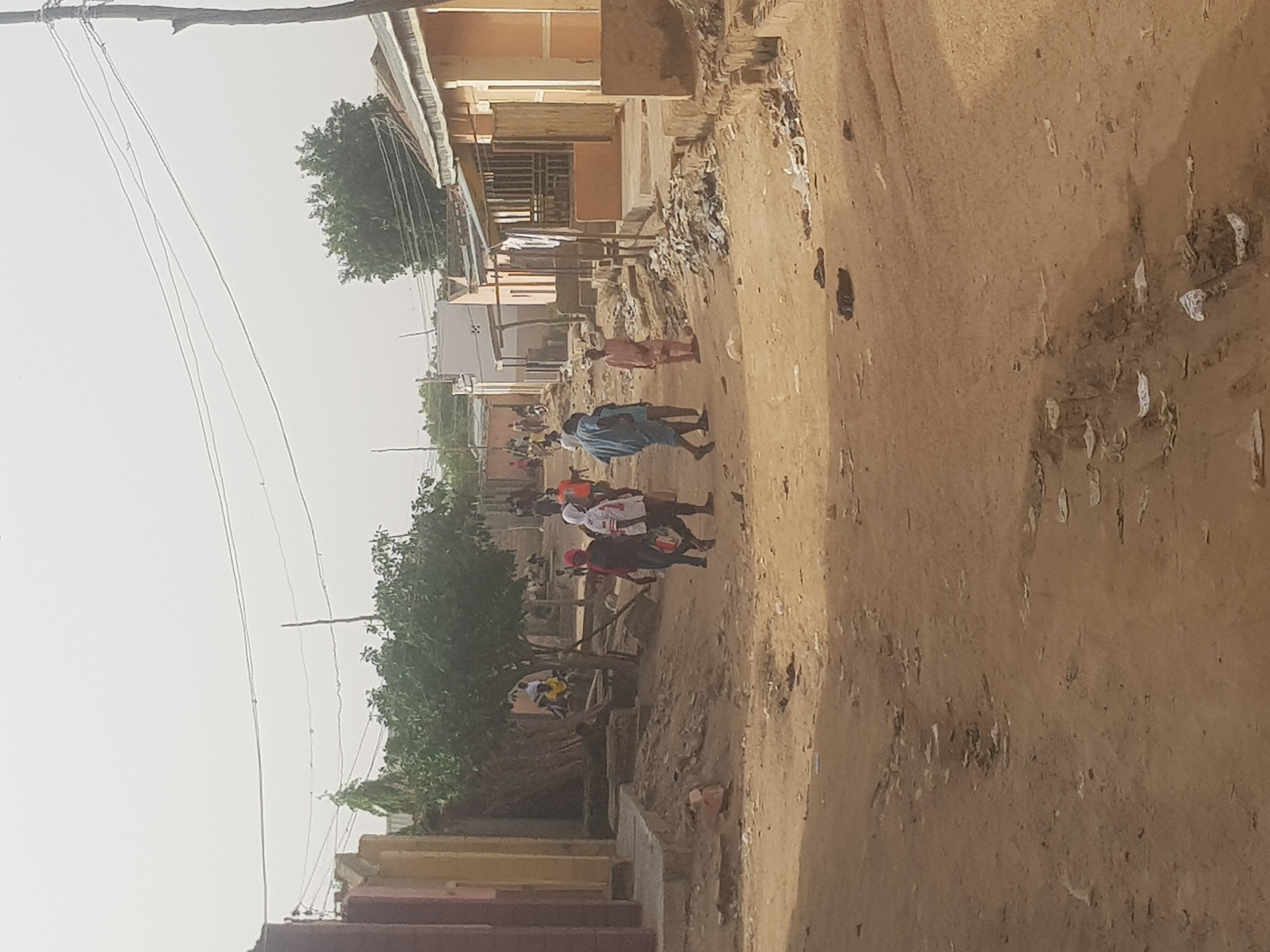 The image depicts a dusty street scene in Dabawa, Nigeria. A group of people are walking along the road. Buildings line the street, and power lines are visible overhead. There is some trash and debris scattered along the side of the road. The ground is dirt with scattered tire tracks and other signs of traffic.