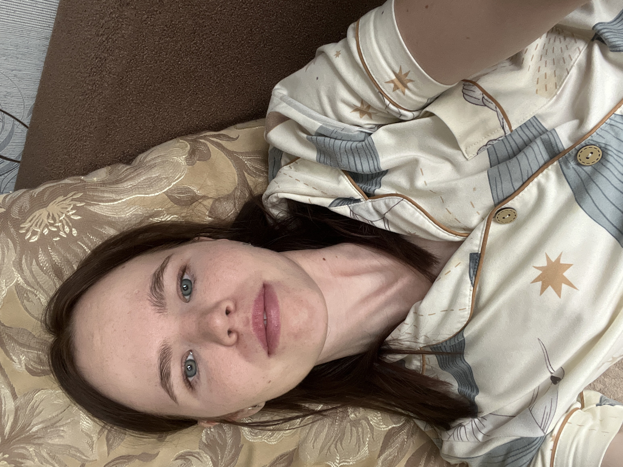 The image presents a close-up portrait of a young woman lying down indoors in Chaikovsky, Russia. She is positioned on her back, looking directly at the camera with a calm, neutral expression. She has fair skin, dark brown hair parted down the middle, and light blue-green eyes. Her upper body is visible, dressed in light-colored, long-sleeved pajamas featuring a printed pattern of stars and abstract designs in muted tones of cream, beige, and light gray, with brown piping and two-holed wooden-look buttons. She is resting on a pillow adorned with a gold and cream floral or damask pattern. In the background, to her left, a brown textured surface, possibly a couch or headboard, is visible, along with a subtly patterned light gray wall. The scene suggests a relaxed, casual moment indoors. No visible text is present.