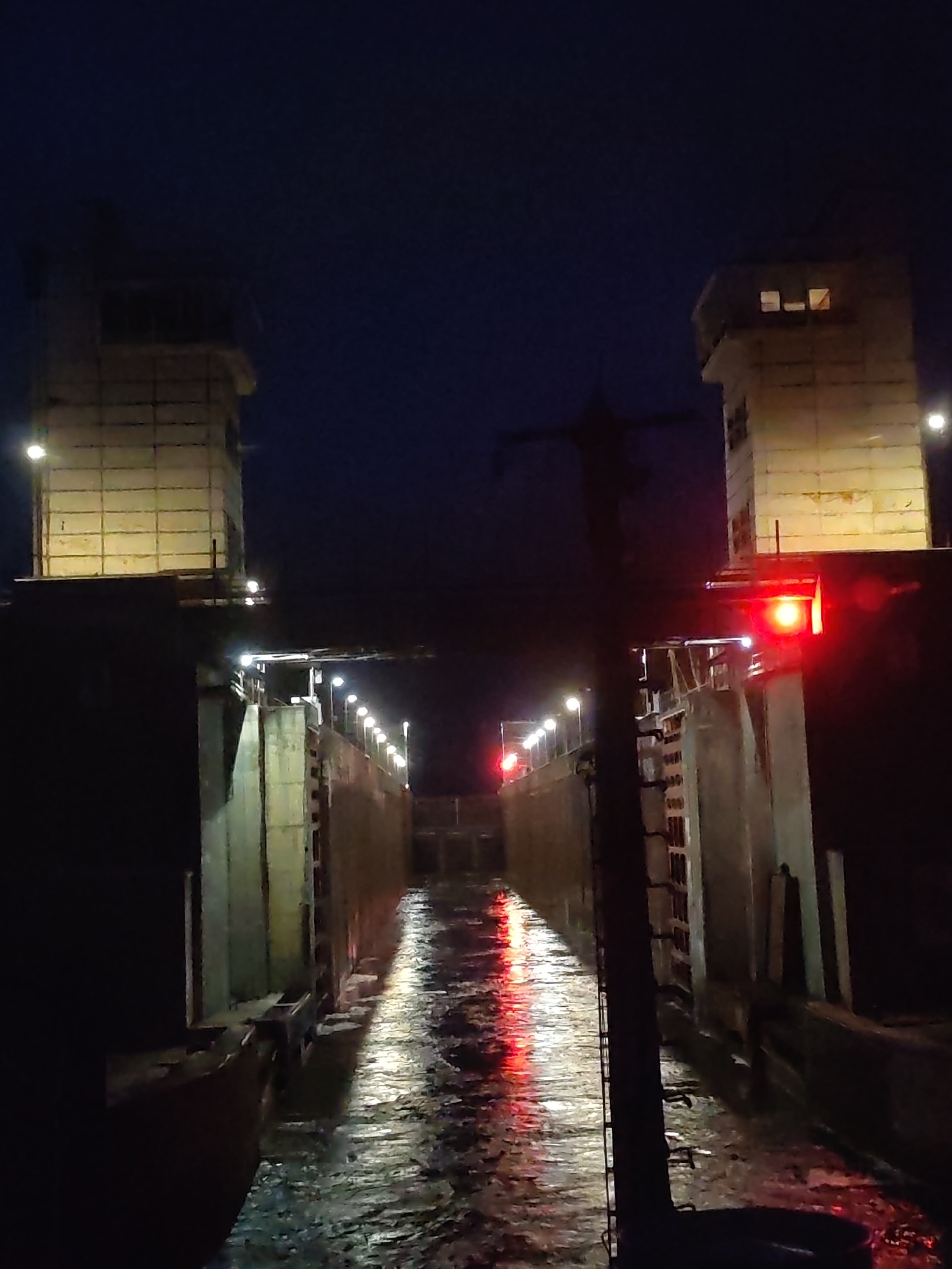 The image depicts a canal lock at night. The lock is flanked by two concrete towers illuminated by yellowish lights, one on each side of the canal. A row of lights lines the edge of the canal walls. Red lights are also visible near the towers. The water in the canal reflects the lights, creating a shimmering effect. A dark, silhouetted structure with a ladder is present in the foreground. The sky is dark blue. The location is in the city of Unknown, Unknown.