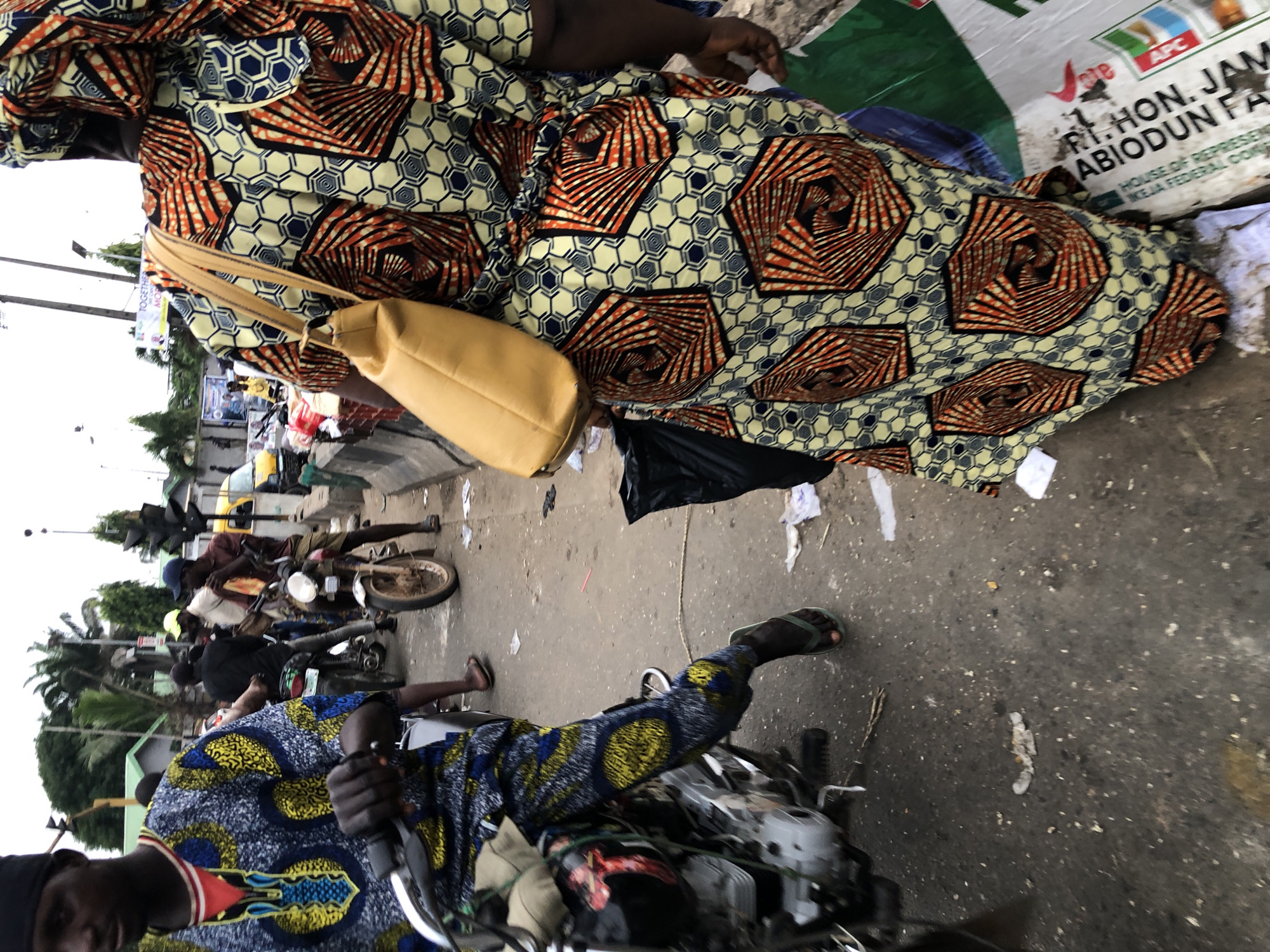 In Ikeja, Nigeria, a group of people are gathered near a road, with several motorcycles visible. In the foreground, a man wearing a blue and yellow patterned dashiki and black cap is seated on a motorcycle, holding the handlebars. To his right, a woman in a brightly colored African print dress and headscarf, carrying a yellow shoulder bag, walks away from the camera. Behind them, other individuals on motorcycles and a yellow car are present. The road appears to be paved and somewhat littered. Traffic lights and palm trees are visible in the background, suggesting an urban setting.