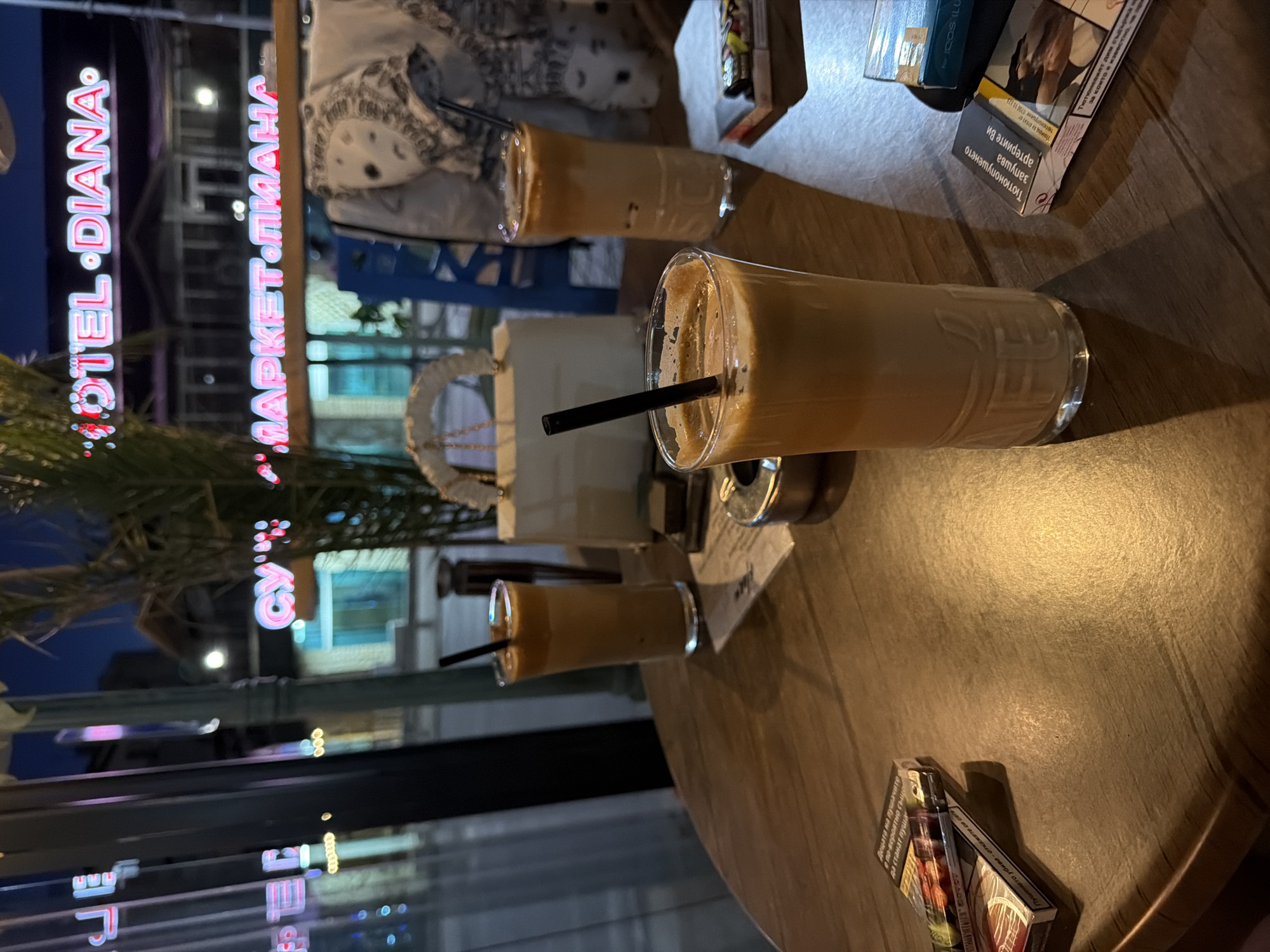 The scene is a cafe or bar in Elhovo, Bulgaria, based on the "Hotel Diana" and "Market Mimama" neon signs visible through a window. The image focuses on a wooden table where three glasses of iced coffee with straws are placed. Next to the drinks are cigarettes and IQOS ILUMA. No people are visible. The image is a close-up, primarily of the table and its contents.