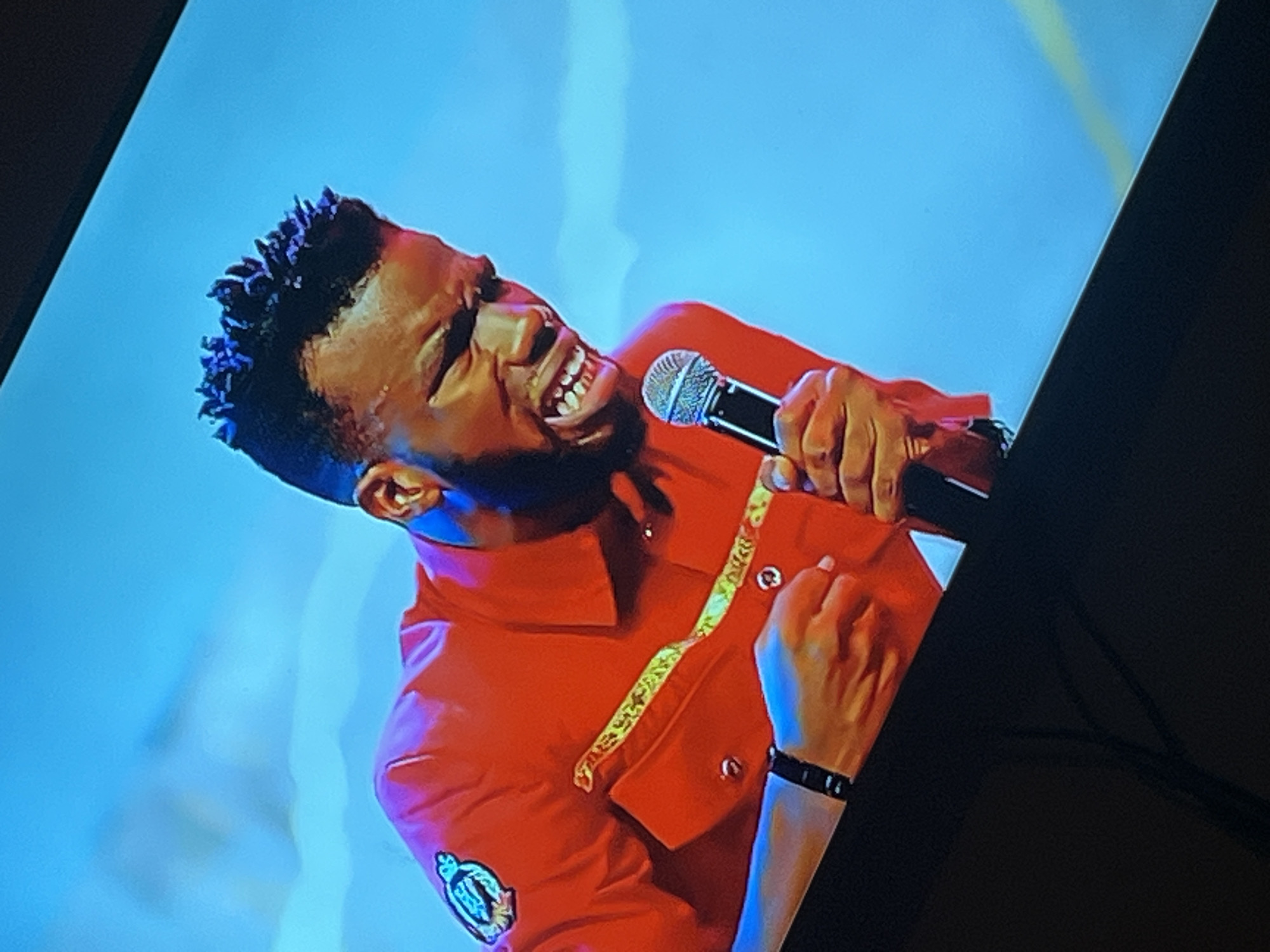 The image shows a close-up of a Black man on a stage, with a bright blue background. He is wearing a red shirt with yellow trim and a patch on the left shoulder. The man is holding a microphone in his right hand and appears to be singing or speaking animatedly, with his mouth open and eyes squinted. His hair is styled in short, spiky dreadlocks. The man is looking downwards and to his right. The lighting is bright and appears to be from stage lights, creating highlights and shadows on his face and clothing. There is a dark area at the bottom of the image, suggesting the presence of a stage or seating area. The man's left arm is visible, with a black wristband on his left wrist. The image quality is slightly grainy. There is no indication of the specific location being Gura Top, Nigeria.