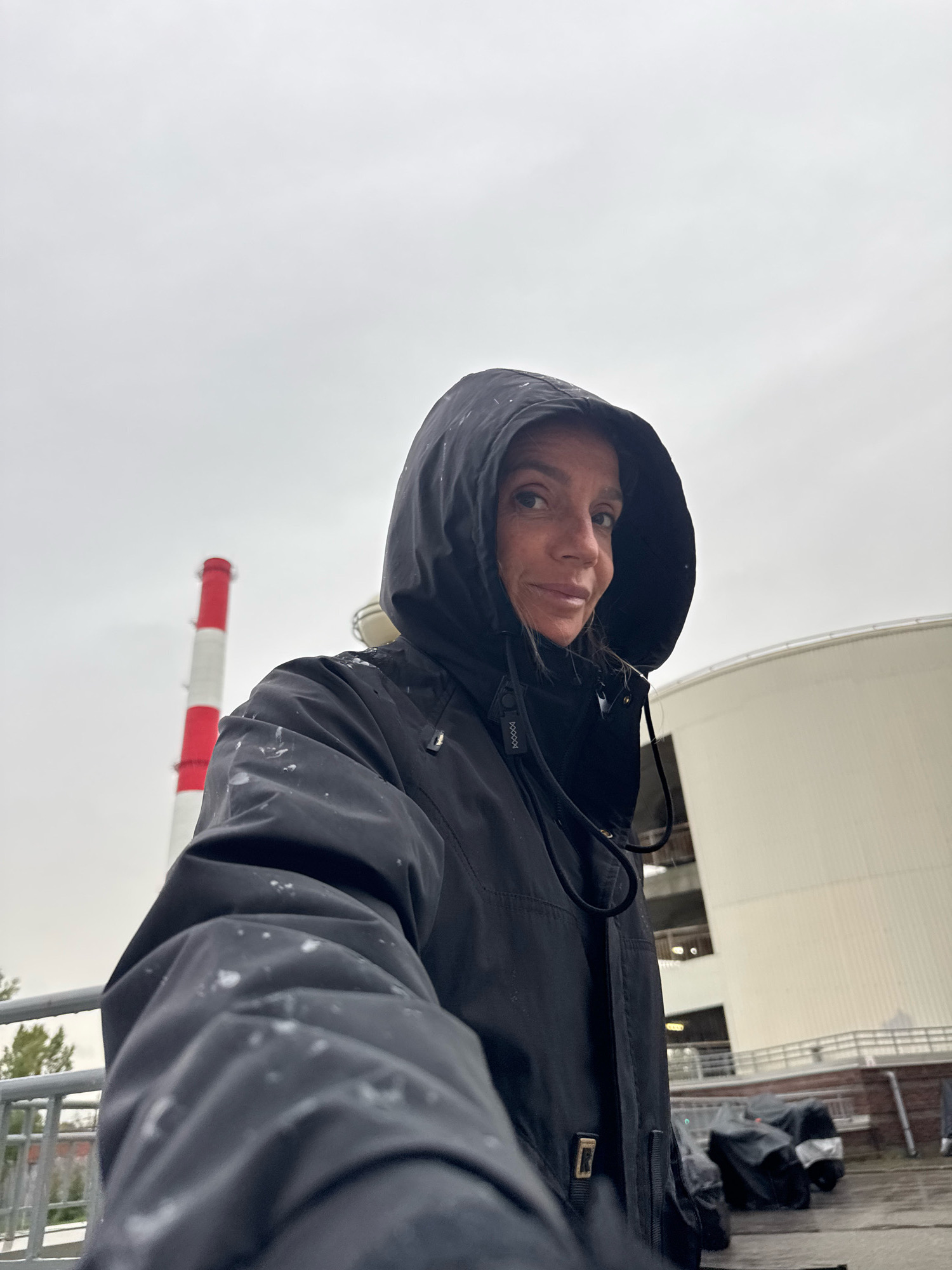 An outdoor, eye-level, selfie-style photograph shows a person wearing a black hooded jacket. The jacket has white splatters on it, and a black drawstring is visible around the hood. The person's face is visible in profile, looking towards the camera. In the background, to the left, is a tall, red and white striped smokestack. To the right of the smokestack is a large, white, curved structure, possibly a storage tank or building. The sky is overcast and gray. There is a metal railing in the foreground and some objects covered with tarps in the background. The environment appears industrial.