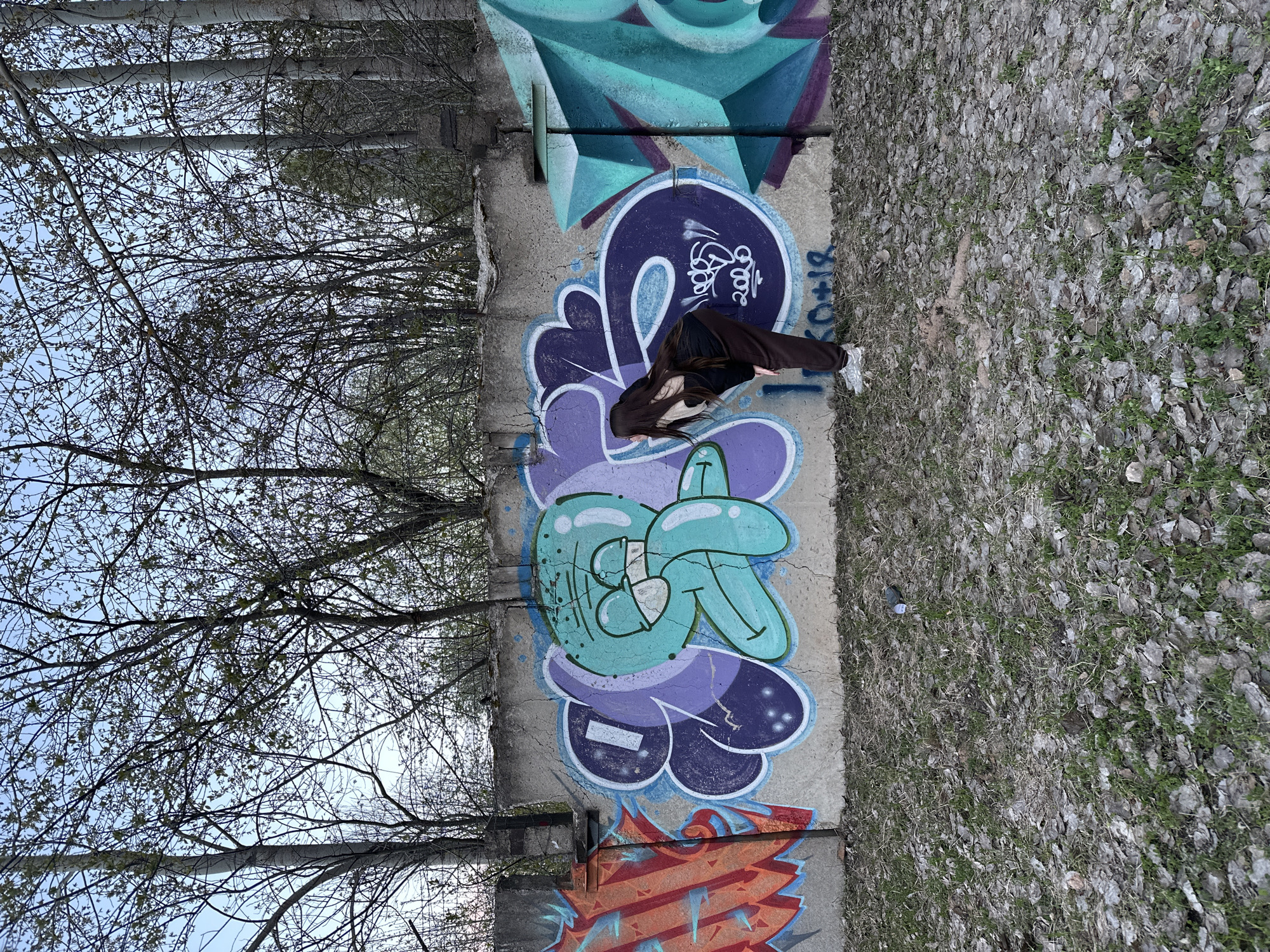A vertical long shot depicts a woman crouched beside a concrete wall covered in graffiti, potentially in Chaikovsky, Russia, based on provided information. She wears a dark brown jacket, black shirt, brown pants, and white sneakers. Her long, dark hair cascades forward as she bows her head.
The graffiti on the wall includes designs in shades of teal, purple, and orange. The ground to the right of the wall is covered in dead leaves and sparse green vegetation. To the left of the wall, there are trees with bare branches, reaching towards the sky. The sky is a pale blue.