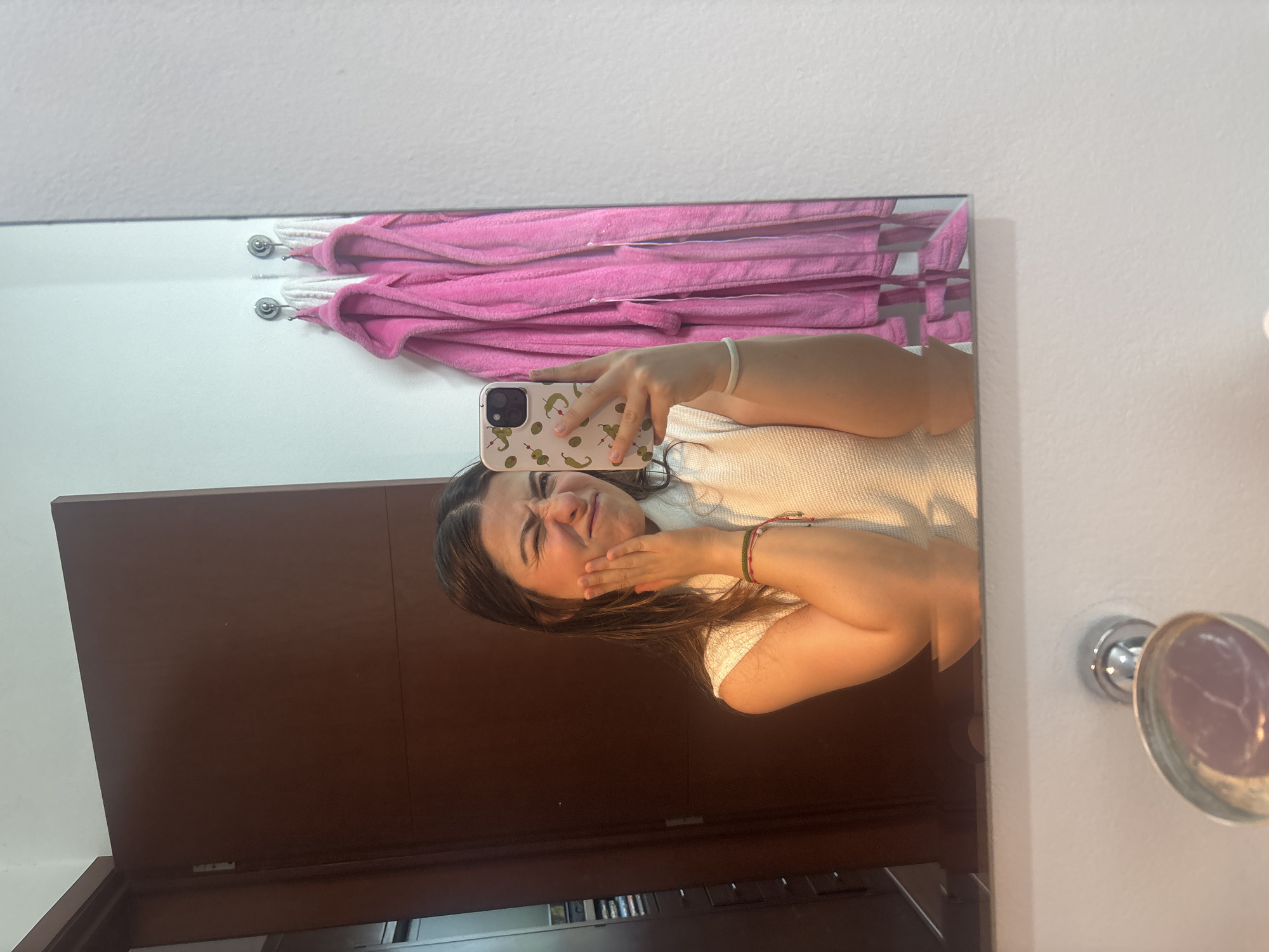 An image, rotated 90 degrees counter-clockwise, depicts a young woman taking a mirror selfie in what appears to be an indoor setting, possibly a bathroom or dressing area in Puebla, Mexico. The woman is the main subject, with dark hair and wearing a light-colored top. Her face is captured in a playful grimace or expression of discomfort, with her eyes squinted and mouth downturned. Her right hand is placed on her cheek or jaw, accentuating the expression. She wears multiple bracelets on her right wrist, some with red and green elements.
She holds a light-colored smartphone with her left hand; the phone features a distinctive case adorned with green olive/pickle and red chili pepper patterns. The phone's camera setup, consistent with an iPhone, is visible. Reflected in the mirror behind her are several pink towels, neatly folded and hung on two chrome hooks against a white wall. To the left of the mirror's reflection, a dark brown wooden door or furniture piece is visible. Outside the mirror on the right, a white wall features a silver, circular fixture with a purple or pinkish reflective surface, possibly a light fixture. No discernible text is visible in the image.