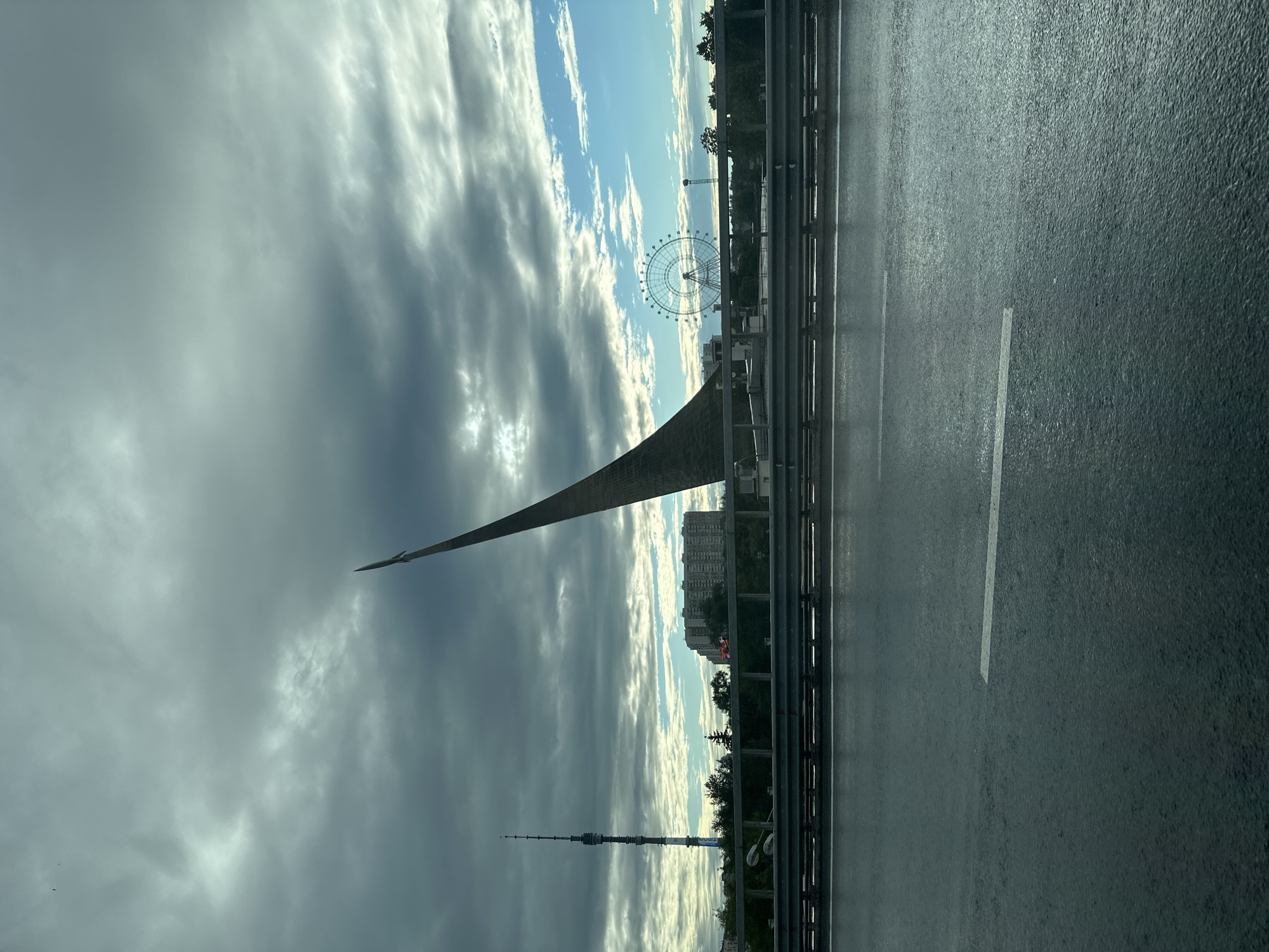 This image captures a view from a road overlooking a cityscape under a dramatic sky. The primary subjects are the Monument to the Conquerors of Space in Moscow, Russia, and the Ostankino Tower, both iconic landmarks. The monument, a tall, sharp, triangular structure pointing upwards, dominates the left side of the frame. In the distance, the Ostankino Tower, a slender, lattice-like structure, pierces the sky. A large Ferris wheel is also visible on the right side of the frame, suggesting an amusement park or entertainment area within the city.
The scene appears to be set during late afternoon or early evening, with the sun casting long shadows and illuminating the clouds from behind. The sky is a mix of dramatic, dark storm clouds and patches of bright blue sky with wispy white clouds, creating a dynamic and somewhat moody atmosphere. The road itself is wet, indicating recent rain or ongoing precipitation, and the surface reflects the sky and surrounding structures. A metal guardrail separates the road from the embankment below. The city skyline features several tall buildings, including residential blocks and other structures, interspersed with trees.
No people are explicitly visible in the image, focusing instead on the architectural and natural elements of the scene. The overall impression is one of urban grandeur set against a powerful sky. The location cues, such as the specific monuments and the general architectural style, firmly place the scene in Moscow. The weather adds a sense of drama and atmospheric beauty to the otherwise static cityscape.