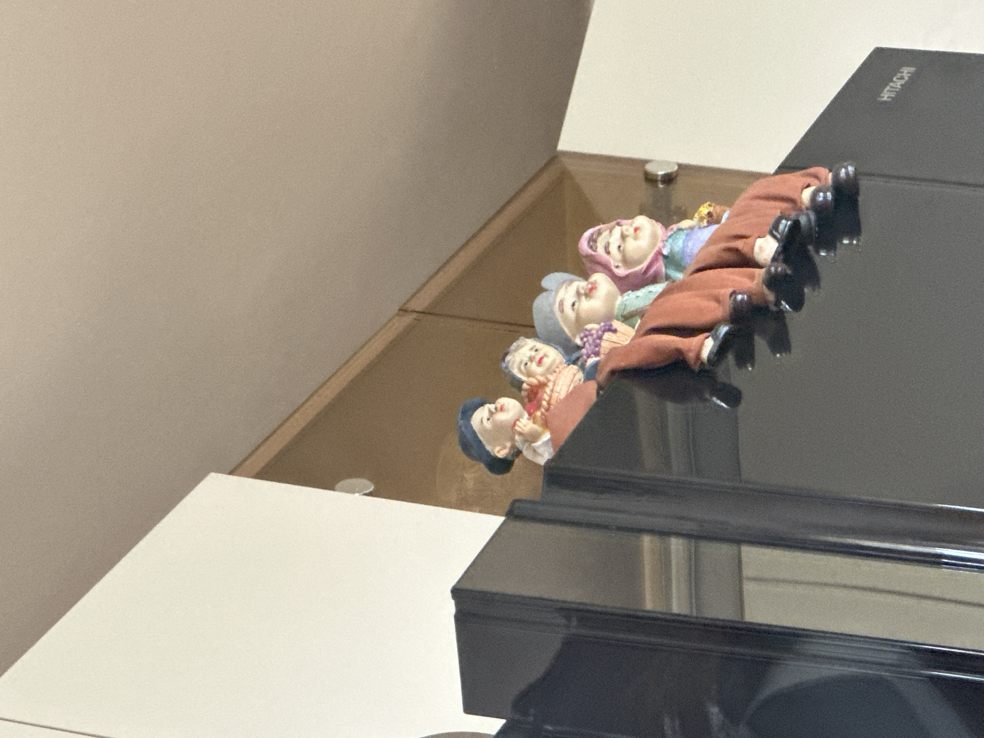 Four small, decorative figurines of elderly women are lying on a dark, reflective surface with the word "HITACHI" visible in the upper right corner. The figurines are dressed in colorful clothing, with two wearing hats. The surface they are on appears to be a part of a piece of furniture with black accents. The background consists of a neutral-colored wall and a white surface. The city and specific location are unknown.