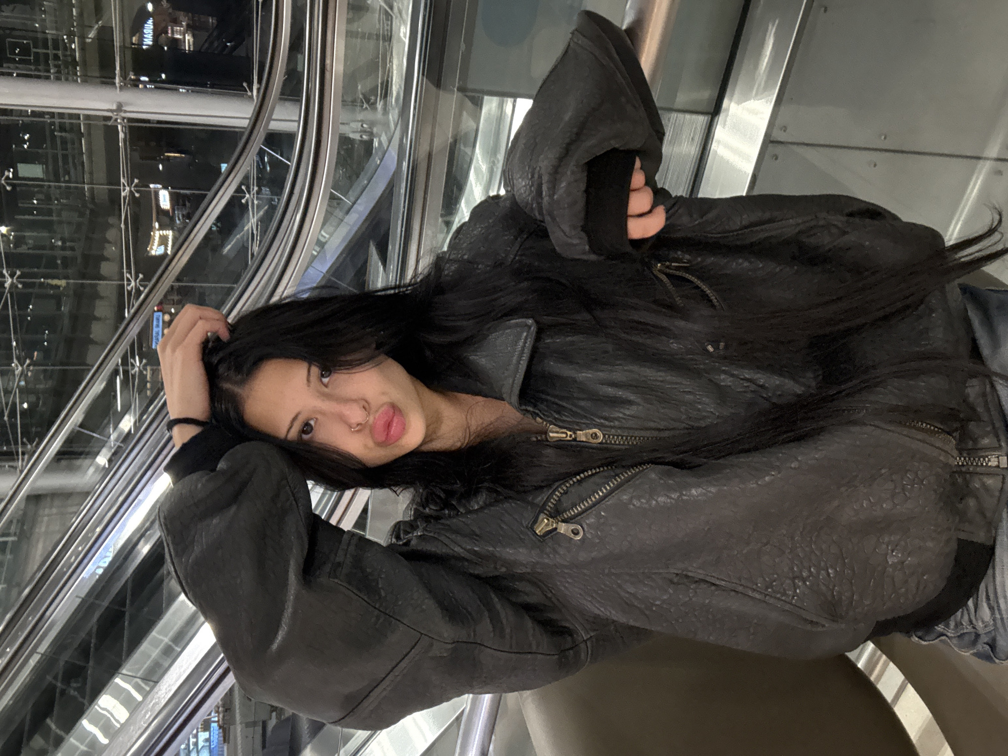 A young woman with long, dark hair and a nose ring is posing for a photo on an escalator. She is wearing a black leather jacket and jeans. Her left arm is raised and her hand is in her hair, while her right arm is resting on the escalator railing. The escalator appears to be in an indoor public space, possibly an airport or shopping mall, with glass railings and reflective surfaces. The lighting is dim, suggesting it is nighttime or the area is poorly lit.