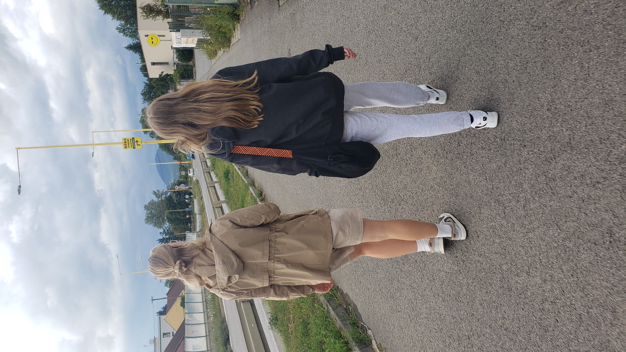 Two individuals, viewed from behind, walk along an asphalt path in Prešov, Slovakia. The person on the left wears a tan hooded jacket, light-colored shorts, white socks, and grey and white sneakers. Their blonde hair is tied in a bun. The person on the right wears a dark grey hooded sweatshirt, light grey sweatpants, and white sneakers. They carry a black bag with an orange and red striped strap over their left shoulder. The path runs alongside a grassy area and a road with a guardrail. In the background, buildings with yellow and white facades are visible, along with trees and a distant hillside under a cloudy sky. Yellow lampposts are positioned along the path. A sign with Slovak text and a parking symbol is mounted on one of the lampposts.