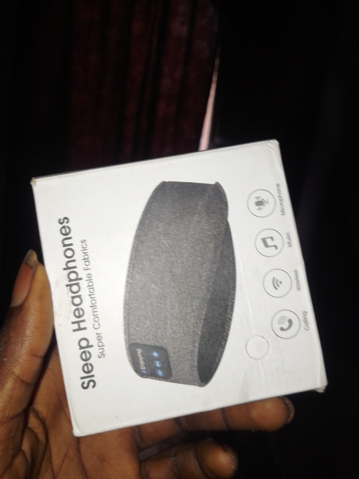 A white box with the words "Sleep Headphones" and "Super Comfortable Fabrics" printed in black is held in a person's hand. The box features graphics of a microphone, a musical note, a wireless signal, and a telephone receiver. A grey, fabric-covered headband, presumably the headphones, is visible on the box. The background is dark, with hints of red drapery. The scene appears to be indoors in a dimly lit environment. No specific location in Serti, Nigeria, can be determined from the image.