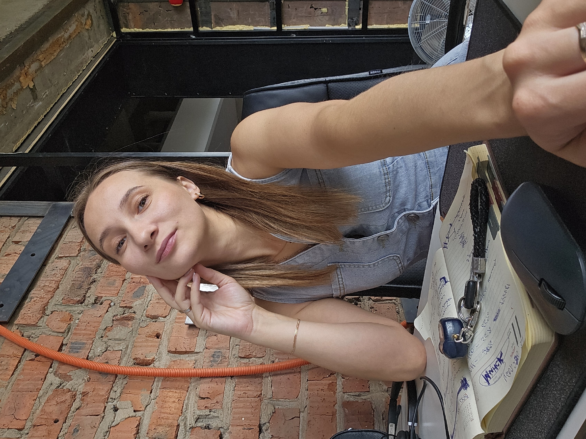 A young woman is taking a selfie while sitting at a desk. She has long, light brown hair and is wearing a denim overall dress. She is leaning her head on her hand and smiling softly at the camera. On the desk in front of her is an open notebook with handwritten notes, a black computer mouse, and headphones. The background shows a brick wall, an orange cable, and a metal fan. The overall impression is casual and relaxed.
The time of day appears to be daytime, possibly late morning or afternoon, judging by the lighting. The location appears to be an indoor space with an industrial or loft-like aesthetic, indicated by the exposed brick and metal structures. The woman's relaxed posture and gentle smile suggest a feeling of contentment or a moment of downtime.
Visible text on the notebook includes what appears to be handwritten notes and possibly some dates or times, though the text is not clear enough to be fully deciphered.