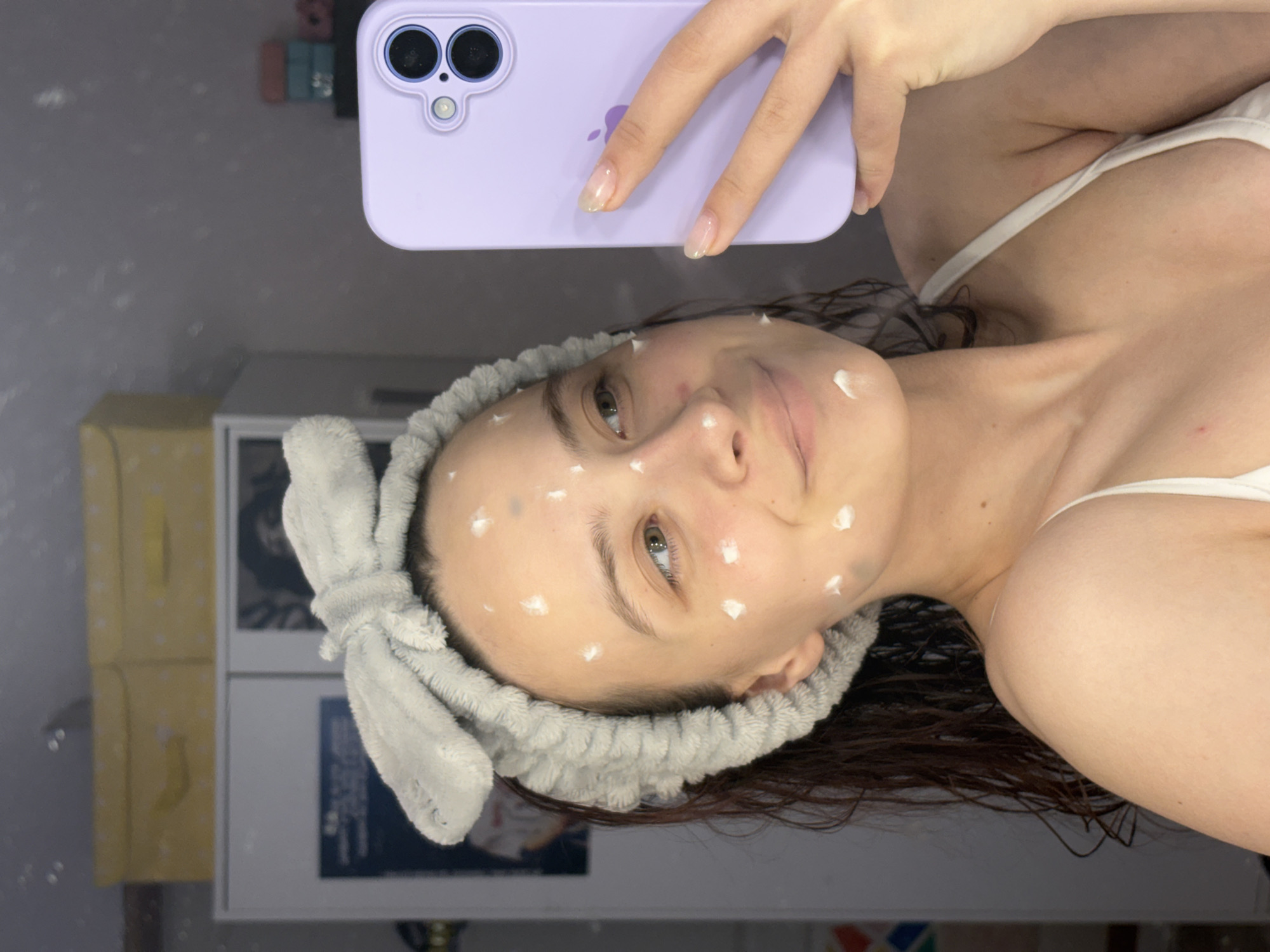 A woman takes a selfie in what appears to be a bathroom setting. The woman is fair-skinned and has dark brown hair and green eyes. She has dots of a white substance applied to her face and is wearing a gray headband with a bow. She is holding a light purple iPhone in her right hand. The background includes a mirror reflecting a neutral-toned wall. A cabinet with visible items is partially visible. The scene is indoors and well-lit. There is no information to indicate the location is in the city of Прикамский, Russia.