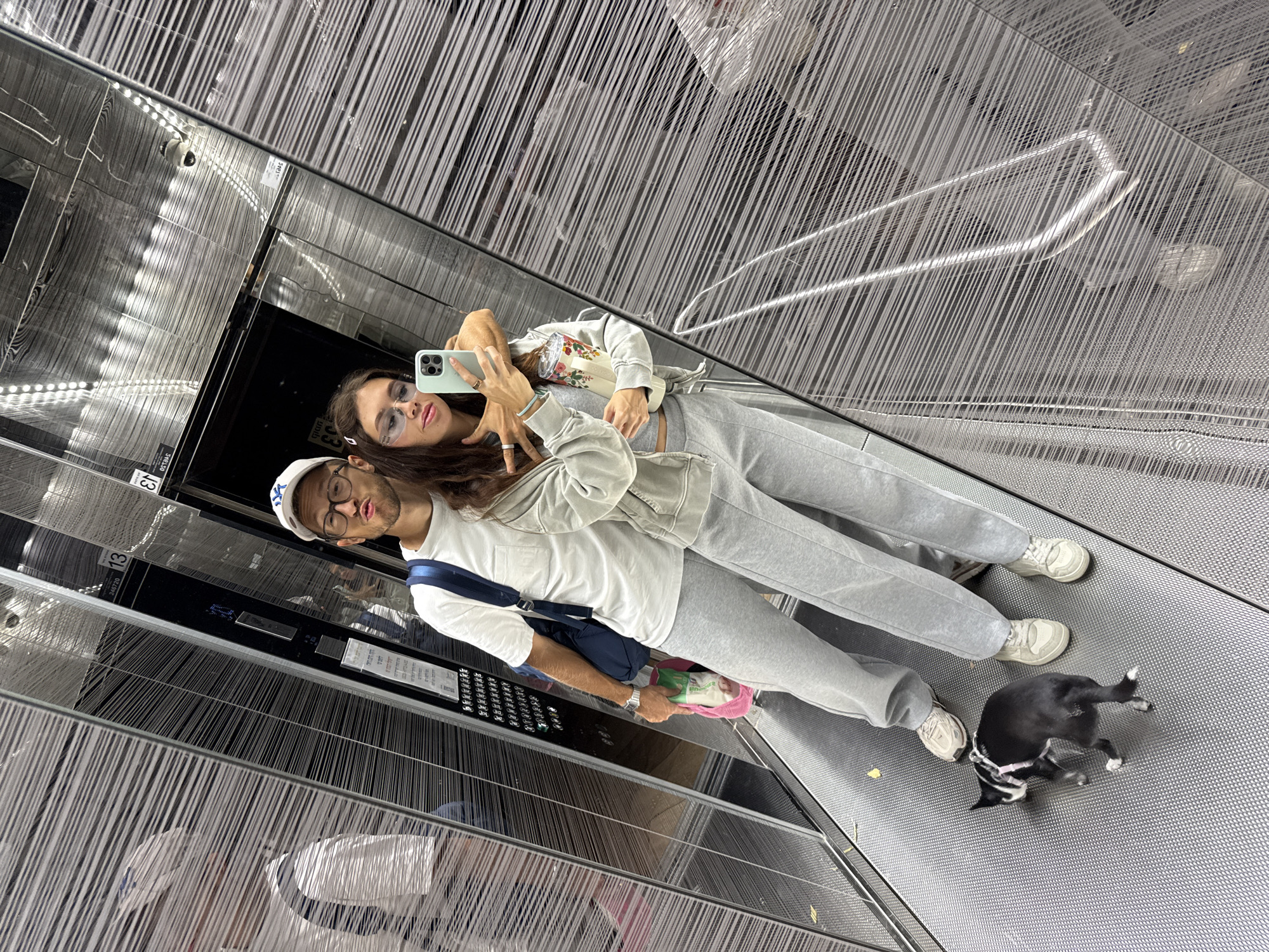 A couple is taking a selfie in an elevator in Tel Aviv, Israel. The woman holds a light green phone, and both individuals are wearing casual clothing. The man has a white hat and a blue shoulder bag. There is a small black and white dog at their feet. The elevator's interior features a metallic, linear design. The location is in Tel Aviv, Israel.