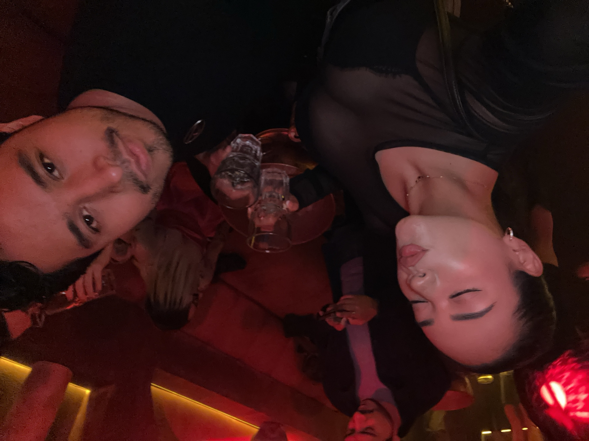 Two individuals pose for a selfie indoors. A man, on the left, looks at the camera with a slight smile. A woman, on the right, has her eyes closed and mouth slightly puckered. They are both dressed in dark clothing. In the foreground, two empty glasses are visible. The lighting appears dim with red tones. Other people are partially visible in the background. The specific location in San Bernardino Tlaxcalancingo, Mexico is not discernible from the image.