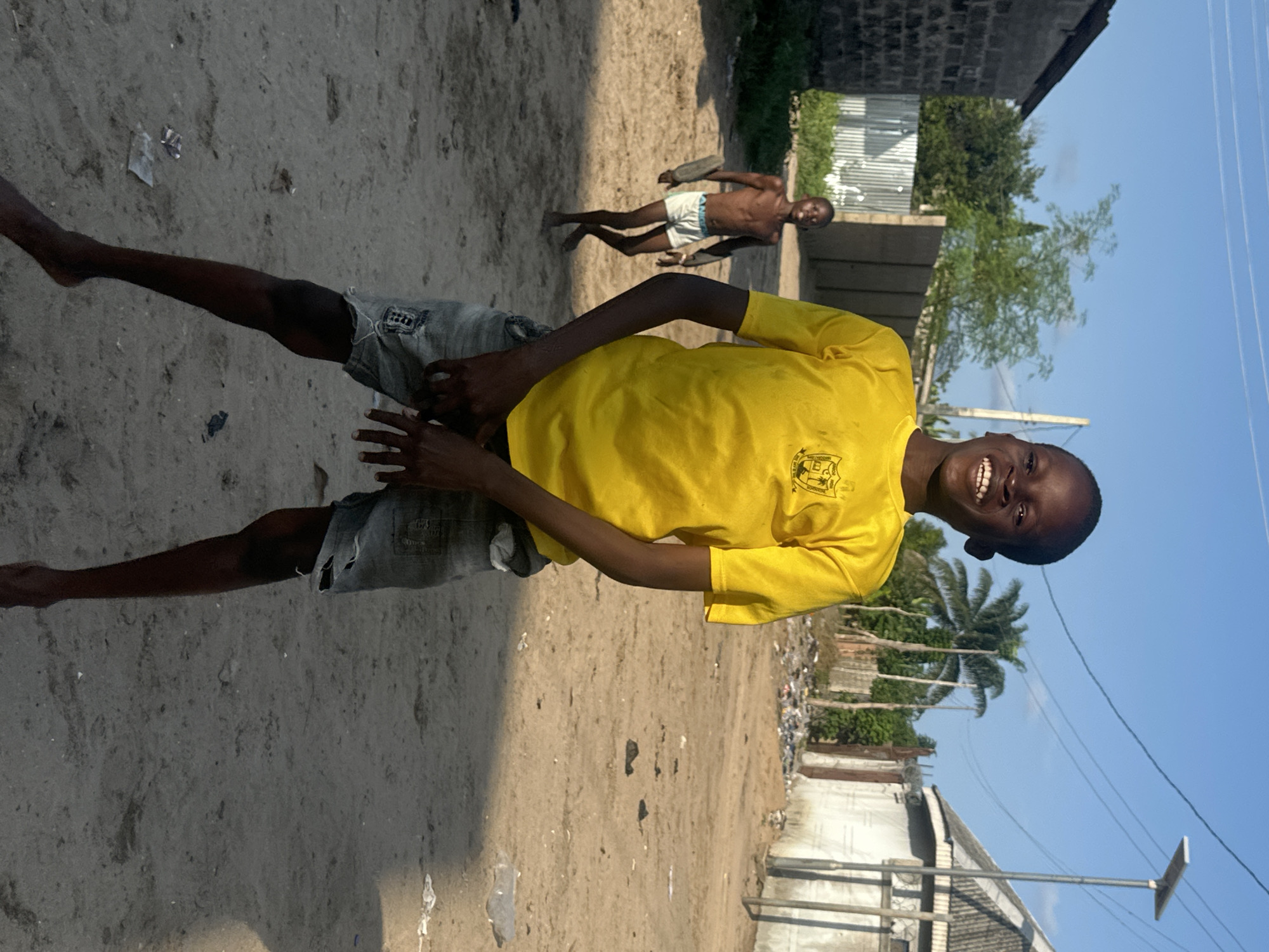 This media file shows two young Black males outdoors on a sunny day. The primary subject, a boy in a bright yellow t-shirt and denim shorts, is positioned in the foreground, facing the camera and smiling broadly. He is standing on a dirt ground. To his left, another boy, shirtless and wearing white shorts, is visible in the mid-ground, facing away from the camera but looking back towards it. He appears to be in motion, possibly running or playing.
The setting is dusty and appears to be a semi-urban or rural area with some buildings in the background. There are scattered debris and some vegetation, including palm trees, suggesting a tropical or sub-tropical environment. Power lines and a street light are also visible, indicating some level of infrastructure. The ground is sandy and uneven. The lighting suggests it is daytime, likely late morning or early afternoon due to the harsh shadows. The overall mood is lively and casual, with the smiling boy conveying a sense of joy and playfulness. There is no visible text in the image.