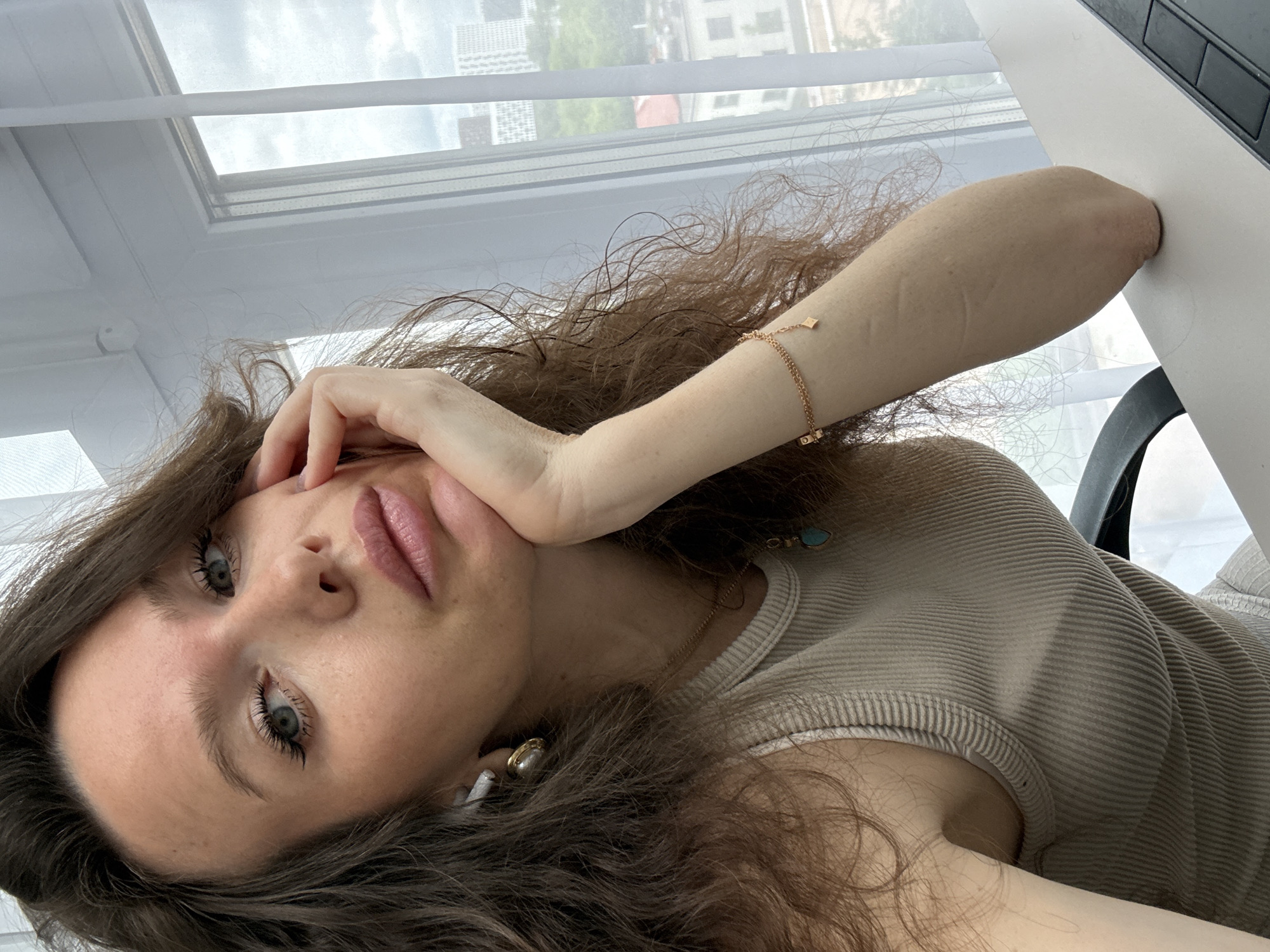 A close-up, angled portrait features a young woman with long, wavy brown hair and bright blue eyes, taken in Khimki, Russia. The image is oriented as if she is lying down or reclining, looking directly at the camera with a neutral to slightly pensive expression, her lips slightly parted.
She has fair skin and is wearing a light-colored, possibly beige or light grey, ribbed tank top. Her left arm is visible, adorned with a delicate gold-colored bracelet featuring small square charms. A gold-colored necklace with a small turquoise or teal pendant hangs around her neck. She has a white earbud in her left ear, partially obscured by a larger, possibly pearl-like earring. Her right hand is gently resting against her cheek and chin.
The background shows a large window with white blinds or sheer curtains, through which blurred urban or suburban buildings and greenery are visible under bright, natural daylight. Part of a white desk or table and a dark chair are visible on the right side of the frame. There is no visible text in the image.