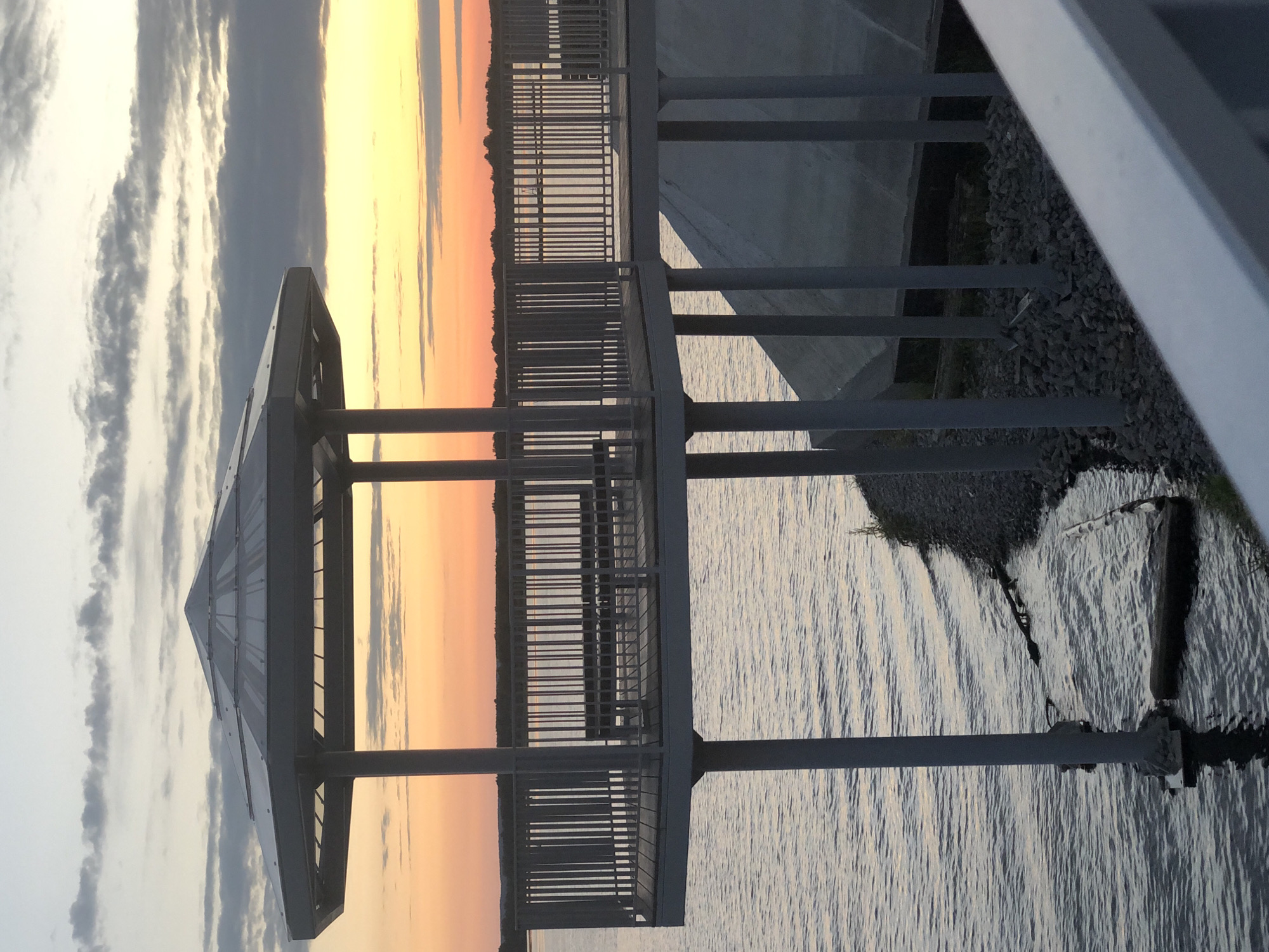 The image shows a waterfront gazebo with a hexagonal roof supported by pillars, situated over a body of water. The gazebo has a wooden deck with a metal railing and benches. In the background, there is a sunset with clouds in a sky transitioning from orange to yellow. The water appears calm. The shoreline is rocky and lined with sparse vegetation. The gazebo is part of a larger structure, likely a pier or boardwalk, with additional support pillars visible extending towards the right side of the image. The scene is devoid of people. This structure is located at the Kama River embankment in Solikamsk, Russia.