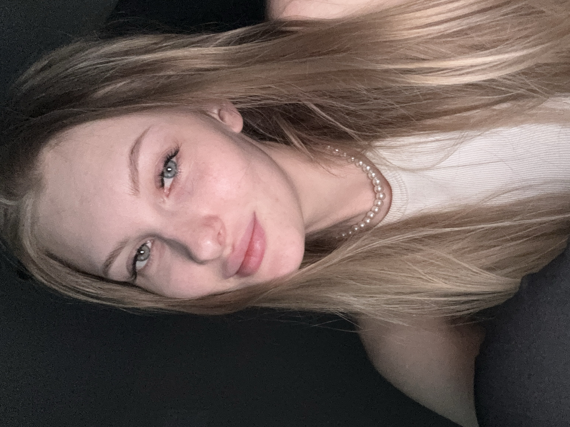 A close-up, rotated portrait features a young woman with long, light blonde hair, reclining in an indoor setting. She has pale skin, light blue or grey eyes looking directly at the camera, and soft pink lips with a gentle, calm expression. Her long, straight hair is spread out around her, contrasting with the dark, undifferentiated background, likely a bed or couch. She wears a light-colored, possibly white or off-white, ribbed top and a delicate pearl necklace. The soft, even lighting enhances her features without harsh shadows, creating a serene and relaxed atmosphere. No text is visible in the image. This media file is contextualized as being from Perm, Russia.