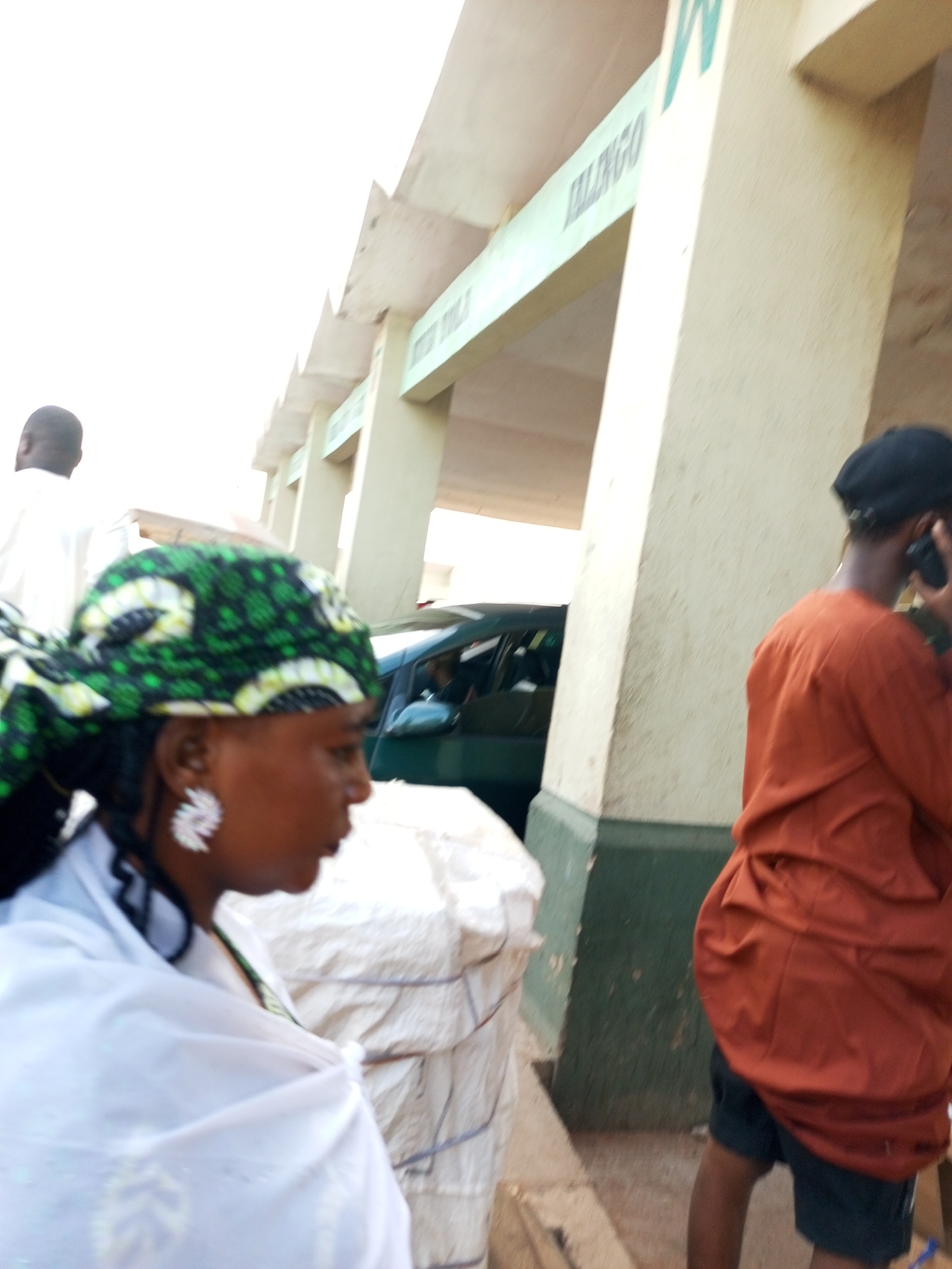 The image captures a scene in Gombe, Nigeria, likely in a market or transportation hub. In the foreground, a woman is visible from the chest up, wearing a green and black patterned headscarf and a white garment. She has a large, ornate white earring in her left ear. Beside her, to the right, is a person from the back, wearing an orange top and dark shorts, speaking on a mobile phone. Behind the woman, a dark green vehicle is parked. Further in the background, there is a building with pillars and signage that appears to read "MALINGO". Another person, a man wearing a white garment, is partially visible on the left side of the image, with his back to the camera. The environment suggests an outdoor setting with an overcast sky. The scene appears to be a public space, with people engaged in everyday activities.
