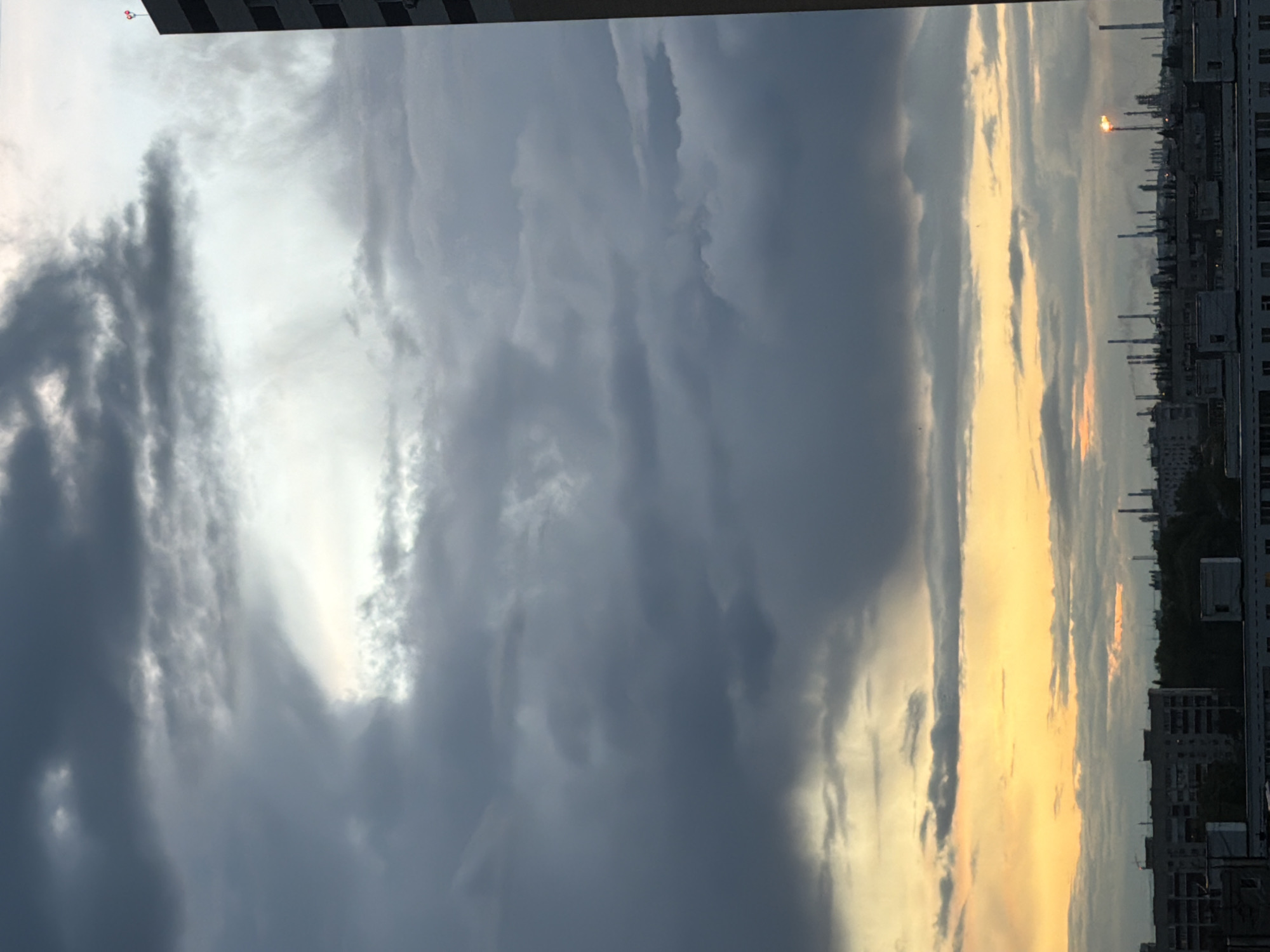 The image displays a dramatic sky over an urban or industrial landscape, rotated 90 degrees clockwise. When viewed correctly, the horizon line runs across the bottom, featuring buildings and industrial structures silhouetted against the sky.
The sky is the dominant feature, characterized by a striking contrast between heavy, dark grey clouds on the left and a brilliant, golden-orange glow on the right. Patches of bright white and light grey clouds are interspersed, suggesting a dynamic weather pattern. The golden light indicates either a sunrise or sunset, with the sun partially obscured by clouds but casting a strong, warm illumination across a section of the sky.
Along the horizon, a row of dark buildings and what appear to be numerous tall, thin industrial chimneys or smokestacks are visible. On the far right, a prominent flare stack emits a bright orange flame or glow, indicating an active industrial site. The buildings are mostly dark silhouettes, showing some window patterns.
The scene captures a moment of dynamic weather and light, likely at dusk or dawn, showcasing the interplay of heavy clouds and warm sunlight over a developed area. No people or visible text are present.