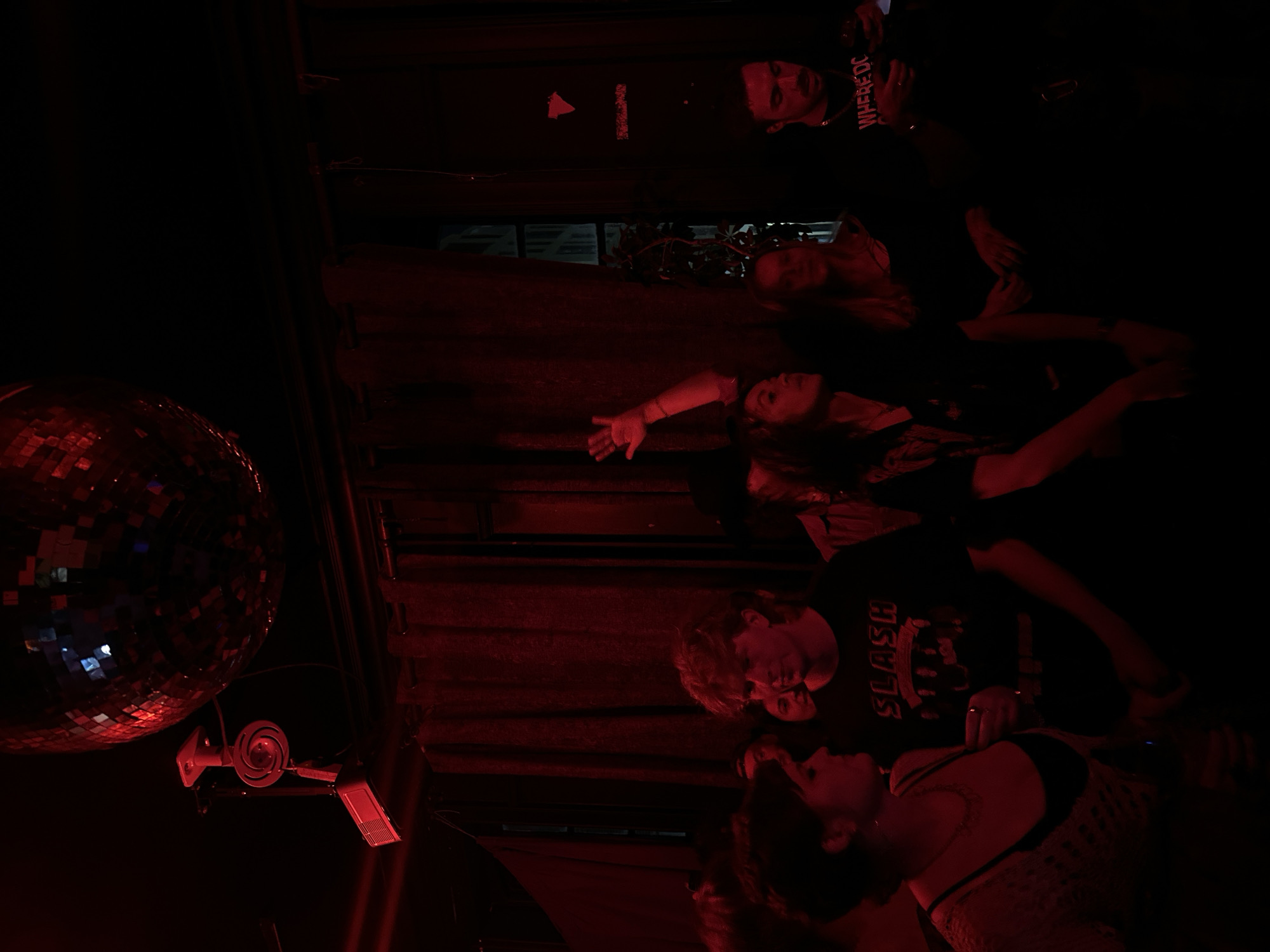 The image captures a dimly lit indoor scene, likely a bar or club in London, bathed entirely in a deep red light. The photograph is oriented vertically, showing a vibrant, nocturnal atmosphere.
In the upper left, a large disco ball hangs, reflecting scattered red light and casting subtle gleams onto the dark surroundings. Below it, a white, spiral-shaped speaker or light fixture is mounted, alongside a rectangular light, both illuminated in red.
Several individuals are visible, relaxed and engaging in social activity. Starting from the top, a man with dark hair and a beard looks towards the right, wearing a dark top with the white text "WHERE'DC" visible. Below him, a woman with lighter hair is partially obscured, looking in the same direction. Further down and to the right, another woman with long dark hair is seen reclining, her left arm outstretched. Her expression is calm.
In the lower portion of the image, two individuals are prominent. A man with light, curly hair wears a dark t-shirt with the word "SLASH" printed in white across the chest. He looks downwards towards a woman next to him, who has dark hair and wears a light-colored, possibly crocheted or mesh top over a dark bra. She wears a ring on her left hand and looks up towards the man, appearing engaged in conversation or interaction.
The setting is dark, with what appears to be dark wooden or curtained walls in the background and a dark upholstered surface where some individuals are reclining. The predominant red lighting creates a moody, intimate, and energetic ambiance typical of an evening event or party. There are no obvious weather cues, but the indoor setting confirms it's a sheltered activity.