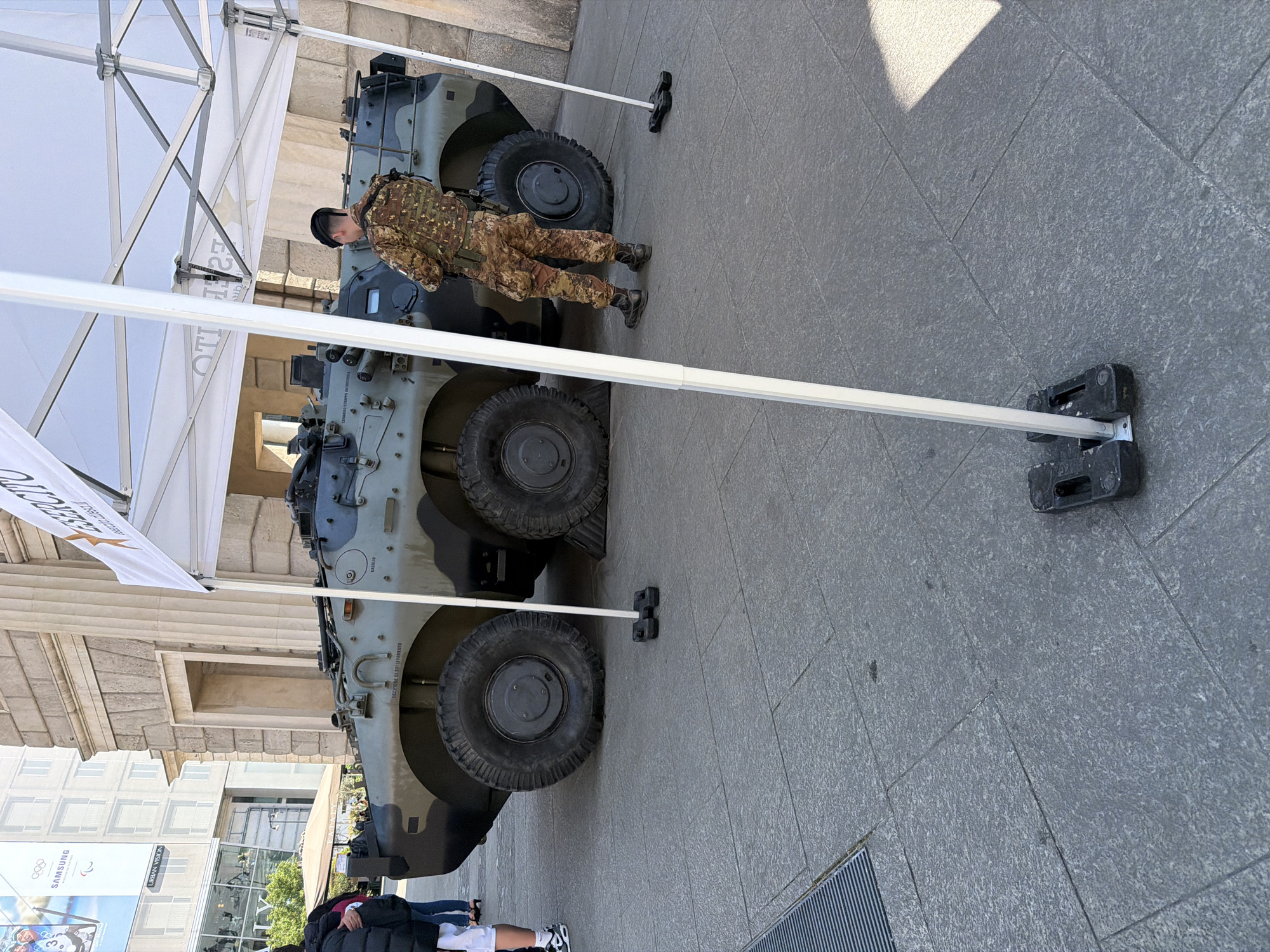 The image shows an outdoor scene in Milan, Italy. A military vehicle, possibly an armored personnel carrier, is prominently displayed under a white tent. A soldier in camouflage uniform stands near the vehicle, appearing to inspect it. The ground is paved with gray stones, and the surrounding environment includes buildings with classical architectural features. In the background, advertisements for the Olympics and Samsung are visible. A few civilians are seen walking nearby, with some near the bottom-left corner of the image. The scene is well-lit, suggesting it's daytime.