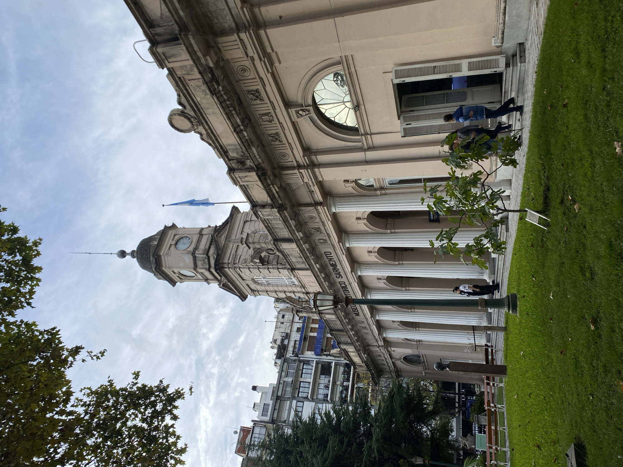 The image shows the Museo Histórico Sarmiento in Buenos Aires, Argentina. The building has a neoclassical facade with a clock tower, columns, and a large arched window. An Argentinian flag flies atop the clock tower. There is a small tree near the entrance. Several people are present; one is sitting on the steps leading into the building and one is sitting on the ground between columns. There is grass in front of the building. A street with other buildings can be seen beyond the museum. The sky is overcast.