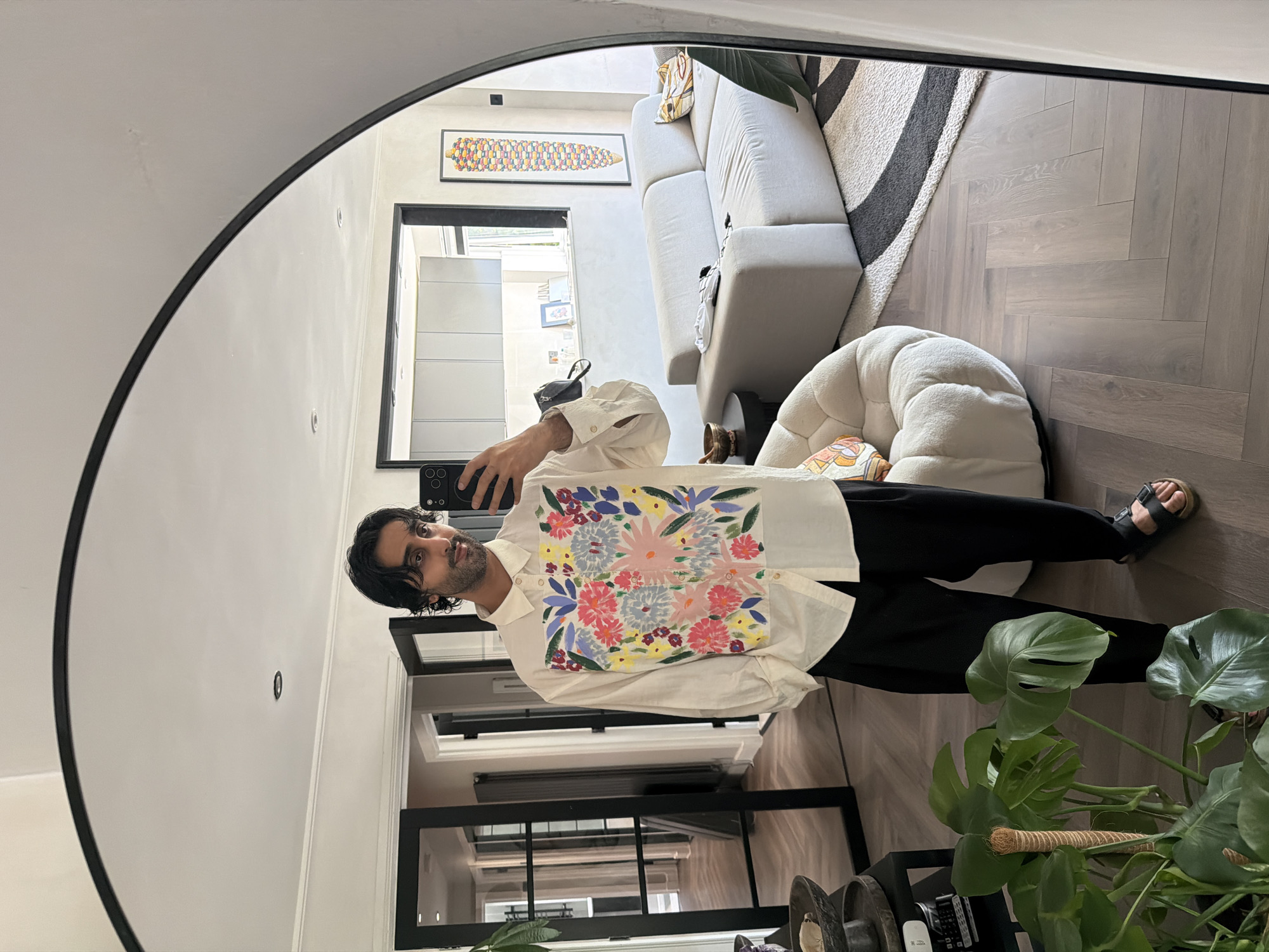 A person is standing in a room and taking a mirror selfie. The person has dark hair and is wearing a white shirt with a floral print on the front and black pants. The person is also wearing black sandals. The room appears to be a living room. There is a couch, a chair, and a rug in the room. There are also several plants. The room has a modern design with hardwood floors. The mirror is large and arched. The image is taken from a slightly low angle. The person is looking at their phone, which is held up to take the photo. The background shows a hallway with glass doors, a kitchen area, and a piece of art on the wall. There is no indication of the specific location being in London, United Kingdom.