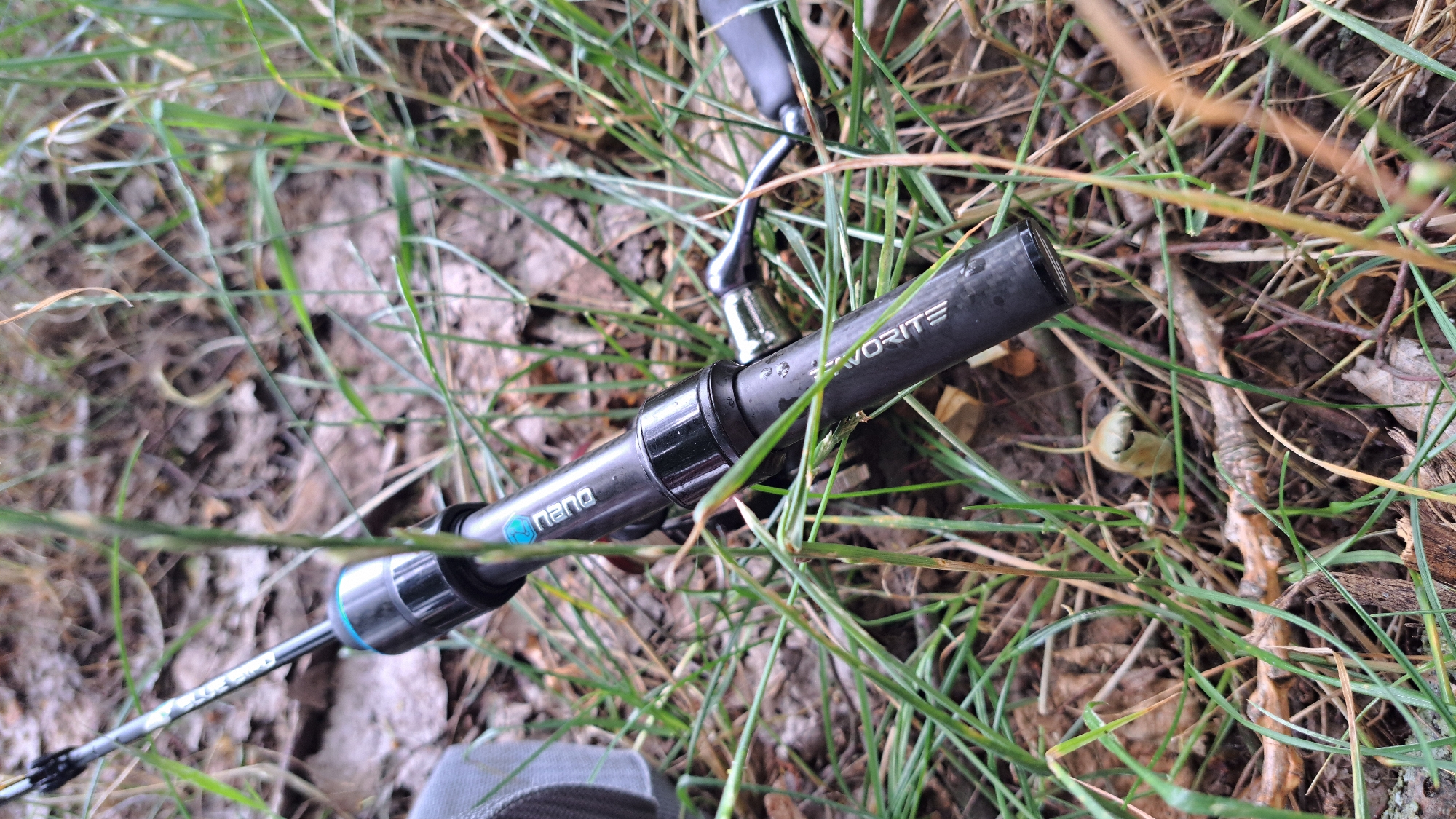 The media file features a close-up, eye-level shot of a fishing rod lying on the ground amidst grass and debris. The rod, primarily dark gray and black with some metallic accents, is the central object. Text visible on the rod reads "FAVORITE" and "nano" with a blue hexagonal logo. The setting appears to be outdoors, likely a natural environment like a field or woodland edge, given the presence of grass, dirt, fallen leaves, and small twigs. The lighting suggests it's daytime, possibly overcast or shaded, as there are no strong shadows. There are no people visible in the frame. The focus is sharp on the fishing rod, with the surrounding environment slightly blurred, emphasizing the object. The overall impression is one of a moment of rest or preparation for fishing in a natural setting. The location context provided is Krosno Odrzańskie, Poland.